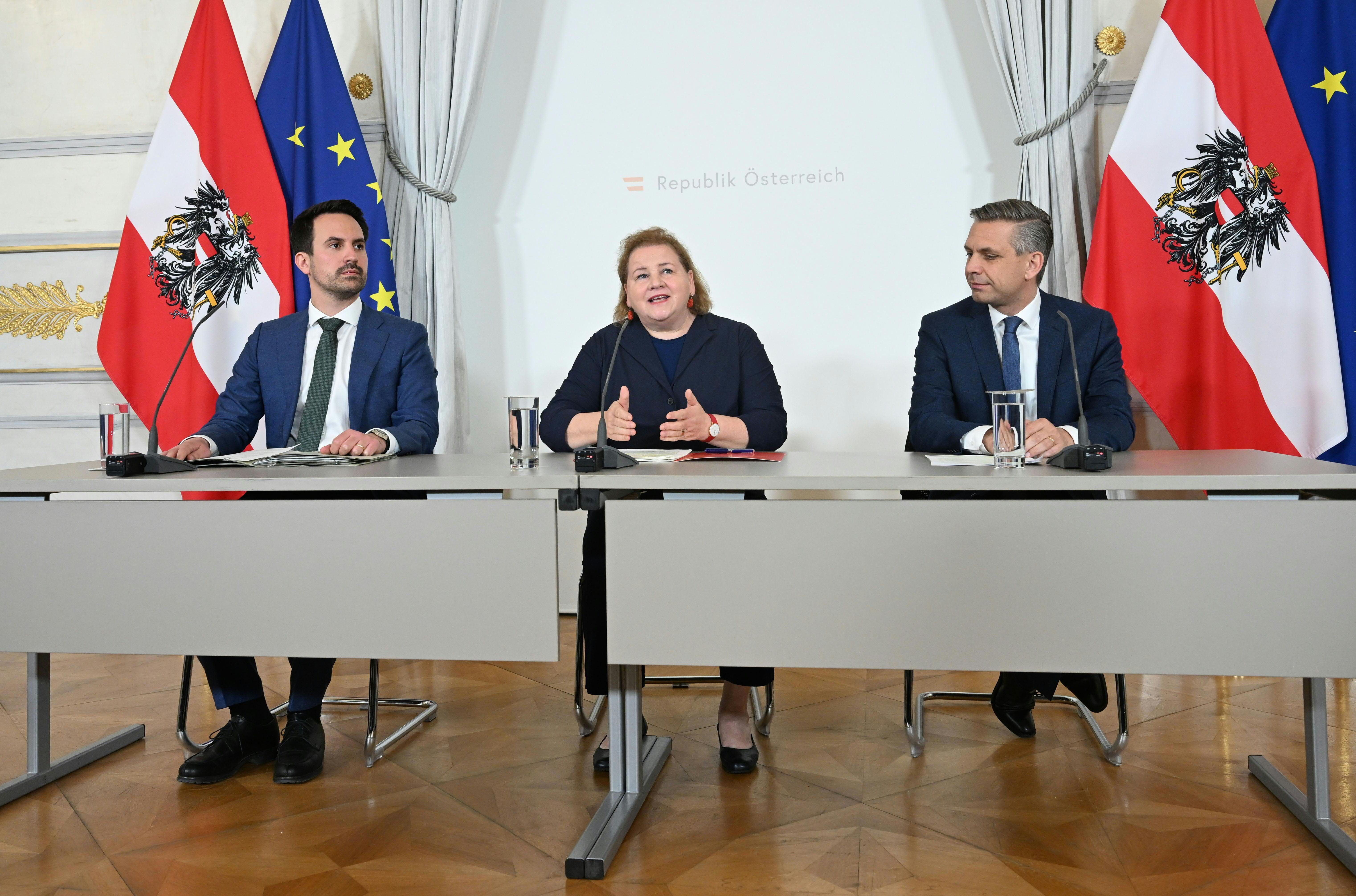 Drei Politiker sitzen im Bundeskanzleramt Wien vor österreichischen und EU-Flaggen bei einer Pressekonferenz.