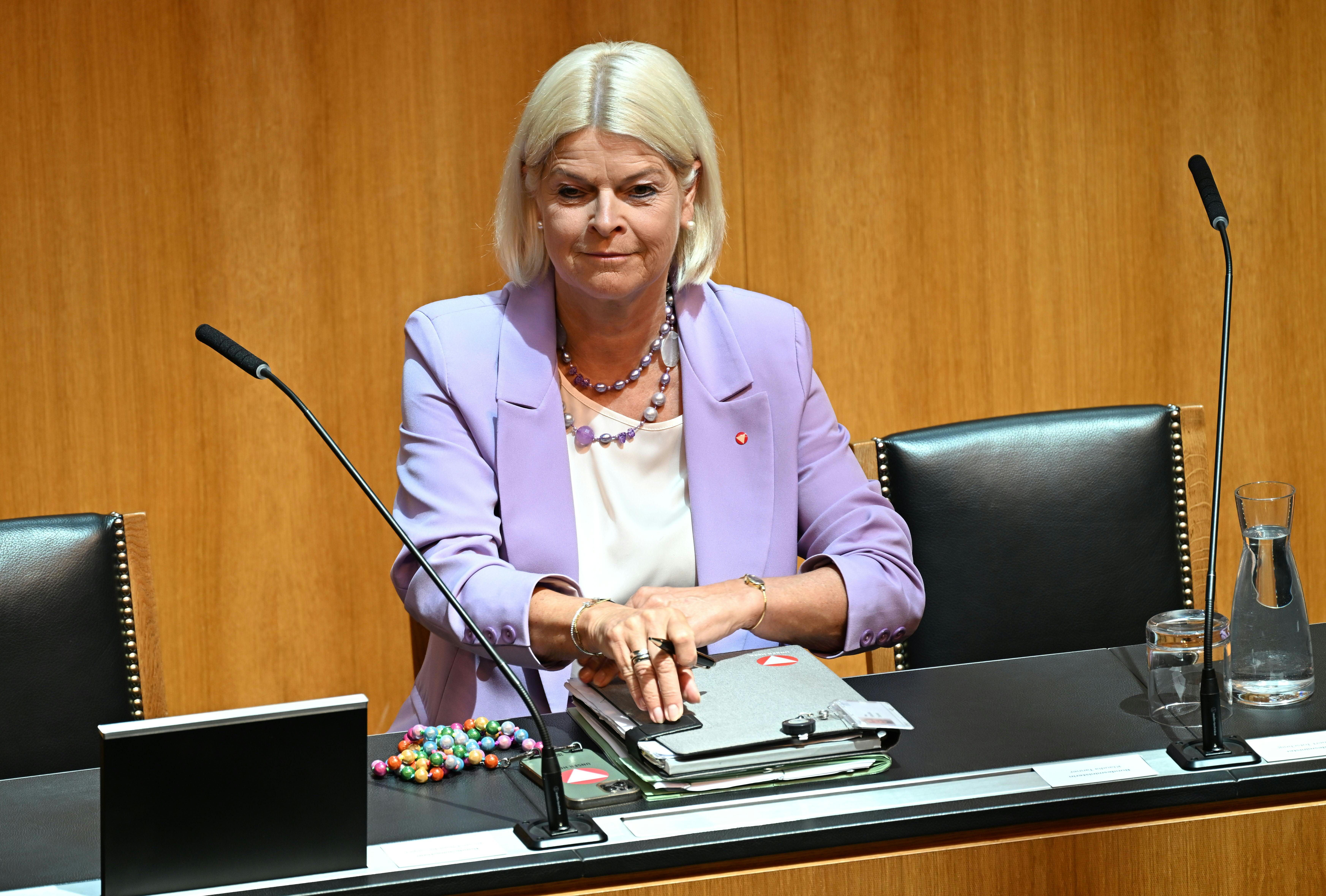 Frau mit hellviolettem Blazer sitzt im Parlament an einem Tisch mit Akten und Mikrofonen.