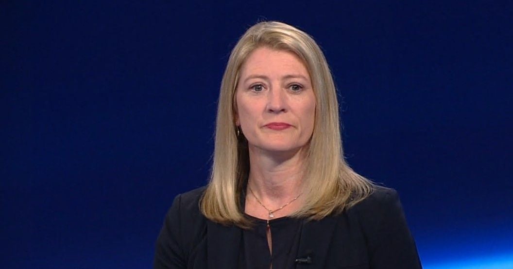 Frau mit blondem Haar und dunklem Blazer vor blauem Hintergrund im Studio.
