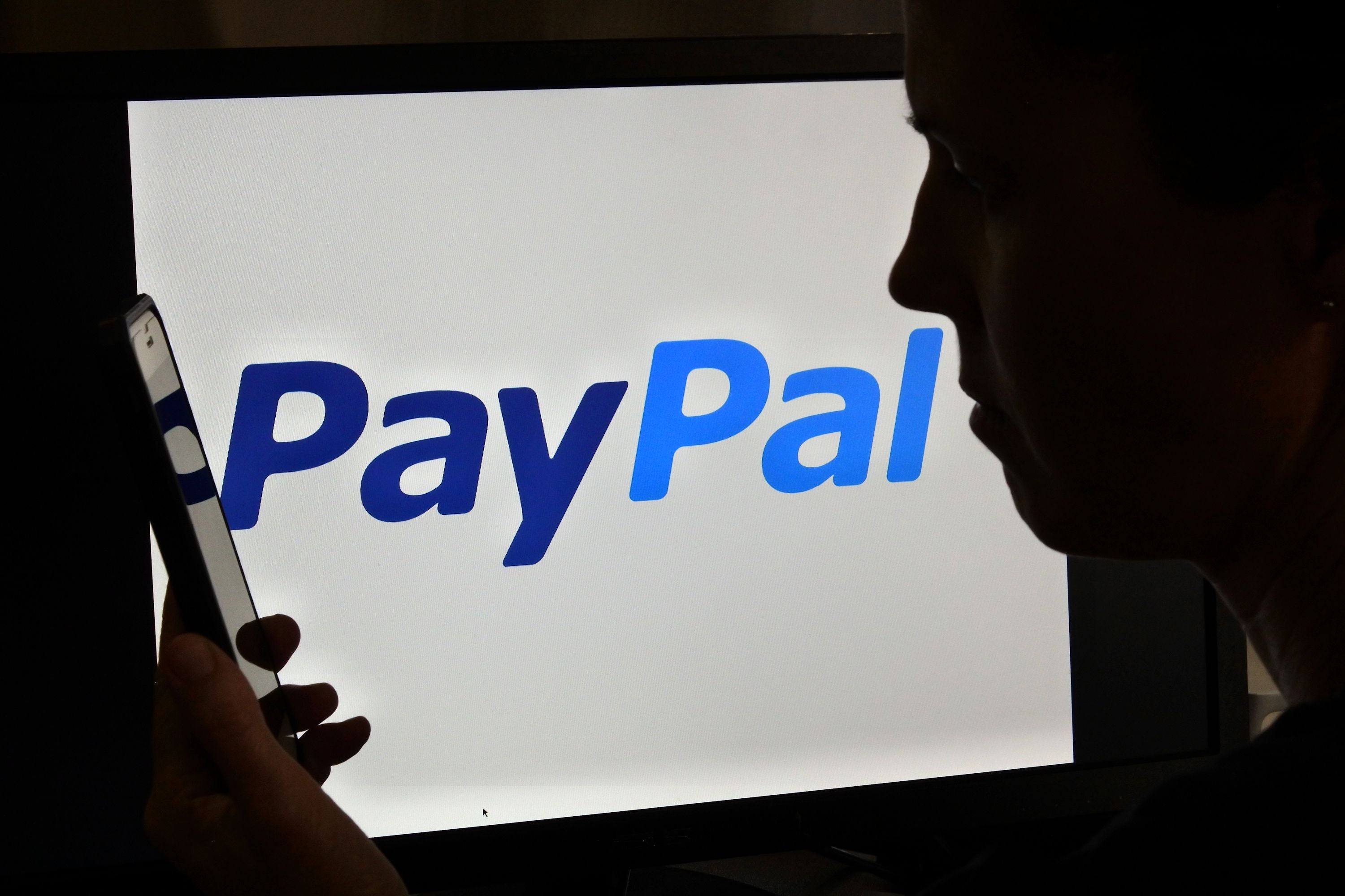 Eine Frau betrachtet ihr Smartphone vor einem Bildschirm mit PayPal-Logo im Hintergrund.