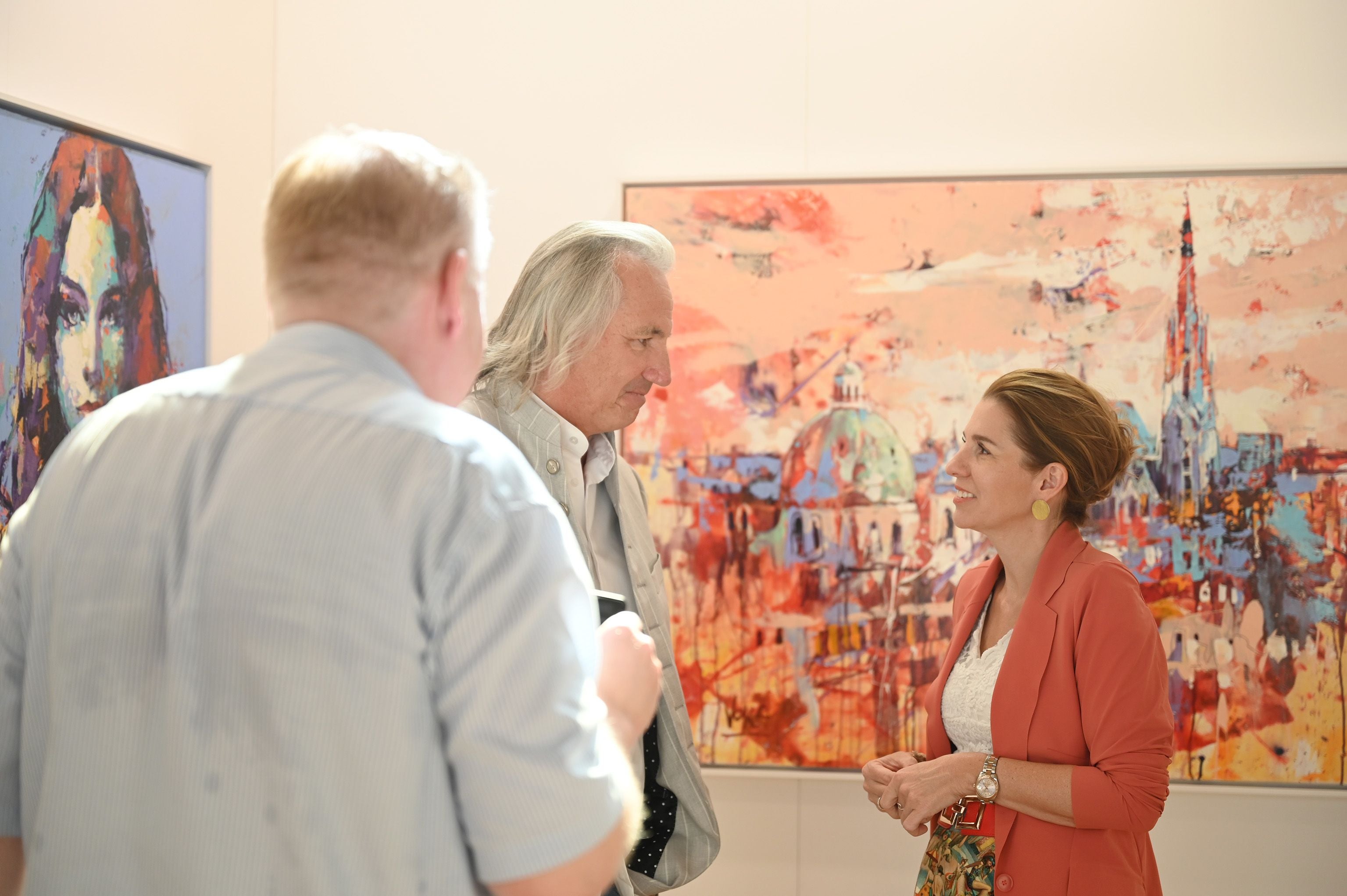 Drei Personen unterhalten sich vor farbenfrohen Gemälden in einer Kunstgalerie.