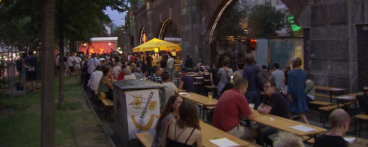 Viele Menschen sitzen und stehen an Bierbänken bei einem Open-Air-Event unter Stadtbahnbögen.