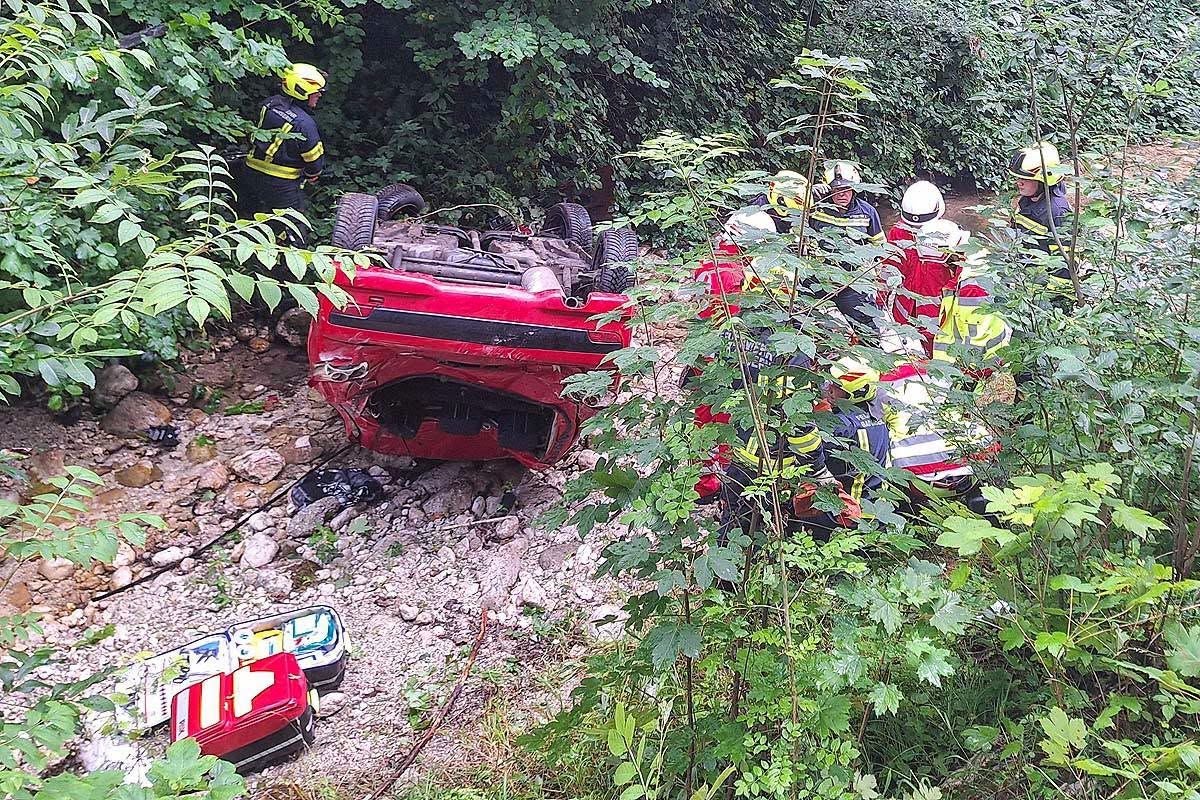 Ein rotes Auto liegt auf dem Dach in einem Graben, Feuerwehrleute leisten Rettungshilfe.