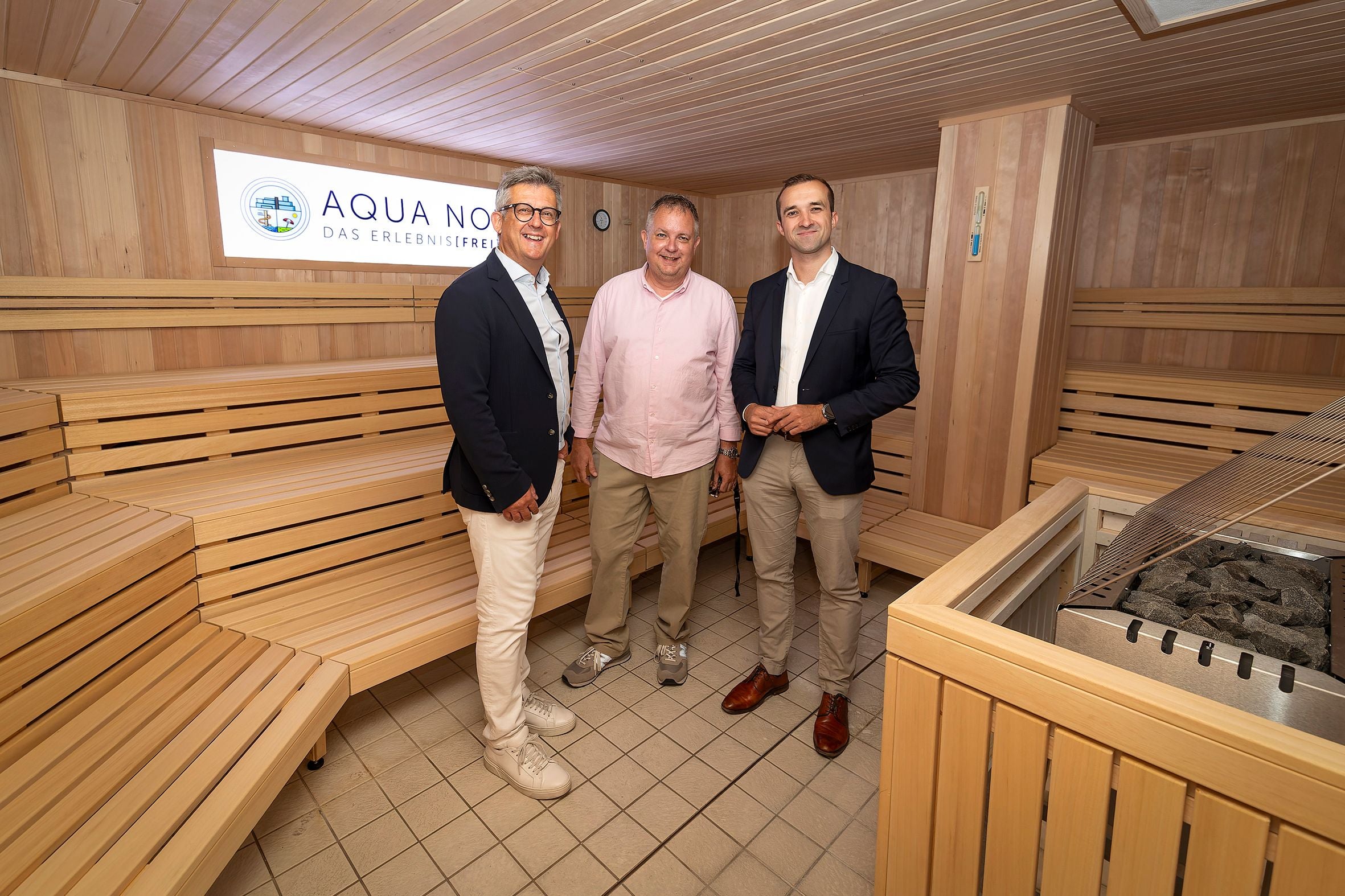 Drei Männer stehen in einer modernisierten Saunakabine mit Holzbänken und Saunaofen.