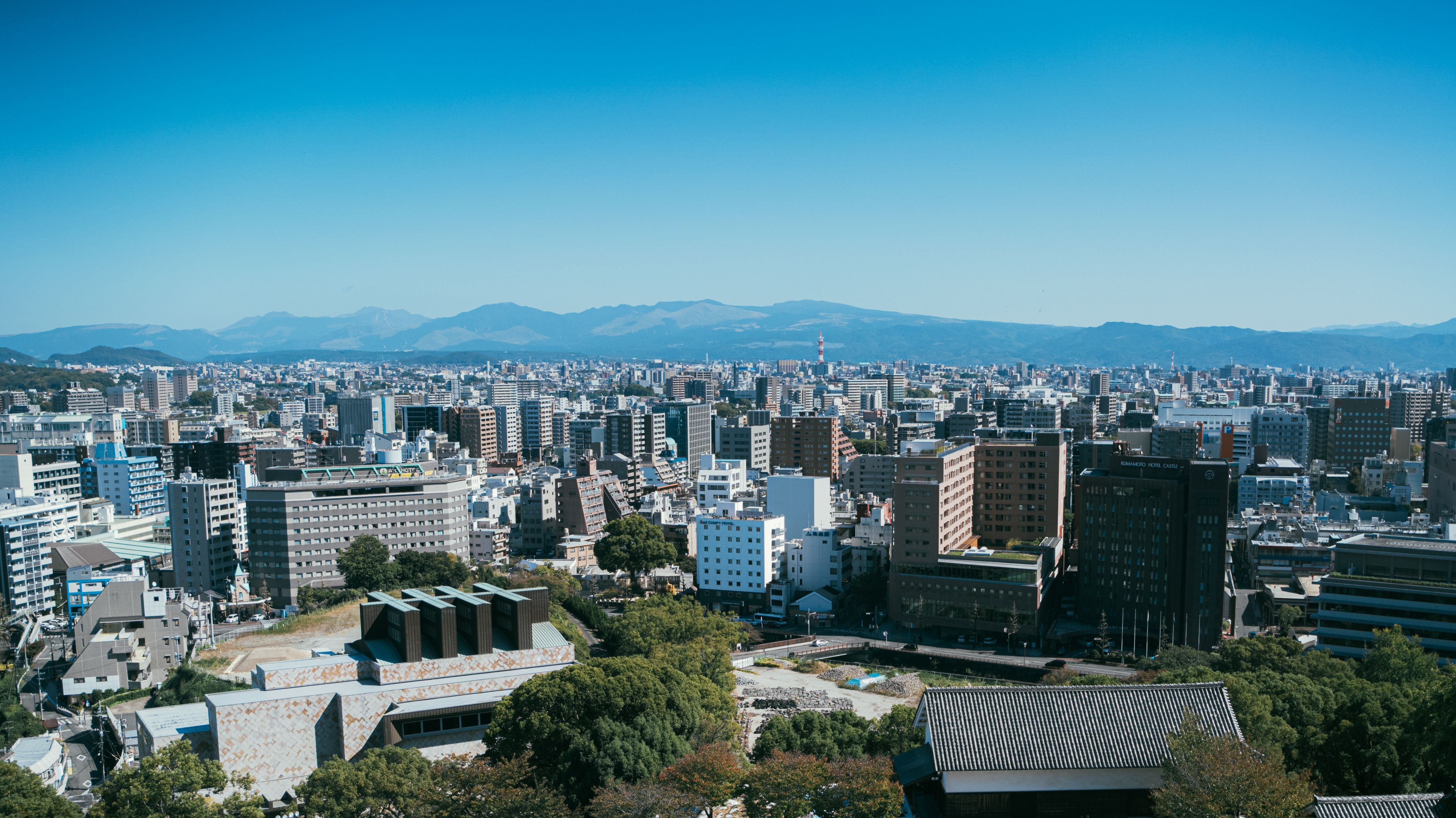 Stadtpanorama von Kumamoto mit vielen Gebäuden und Bergen im Hintergrund bei klarem Himmel.