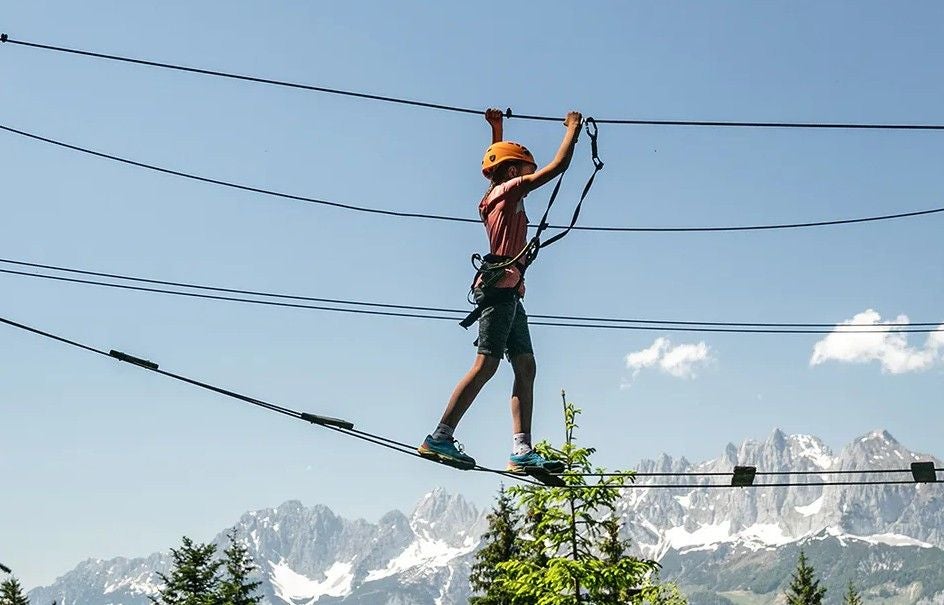 Kind mit Kletterausrüstung balanciert im Hochseilgarten vor Bergkulisse.