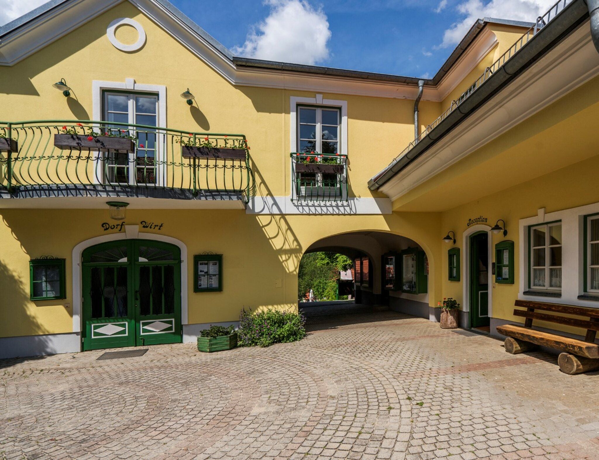 Gelbes Gebäude mit grünem Eingang, Balkon, Bank und Innenhof unter blauem Himmel.