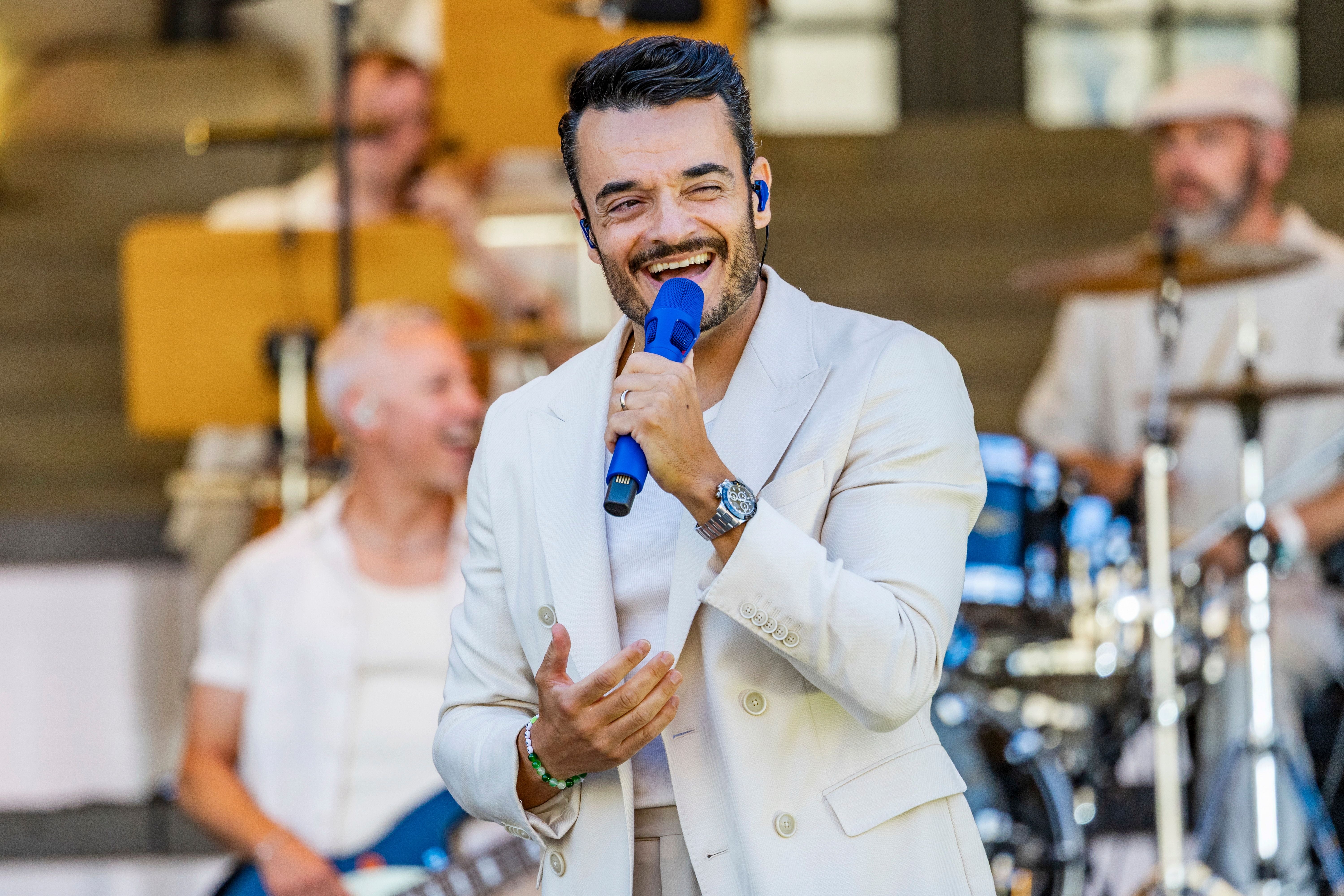 Sänger im weißen Anzug singt mit blauem Mikrofon auf einer Bühne, Musiker im Hintergrund.