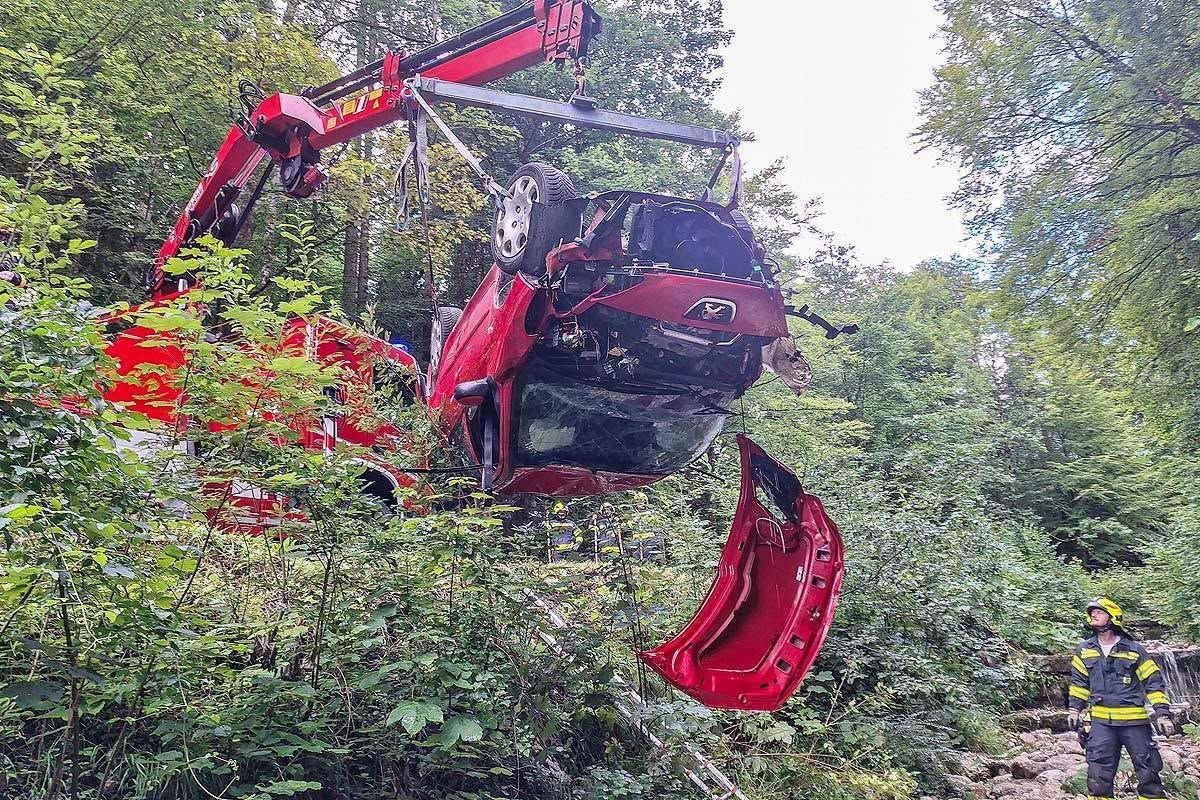 Ein Kran hebt ein schwer beschädigtes rotes Auto aus einem Wald, ein Feuerwehrmann beobachtet.