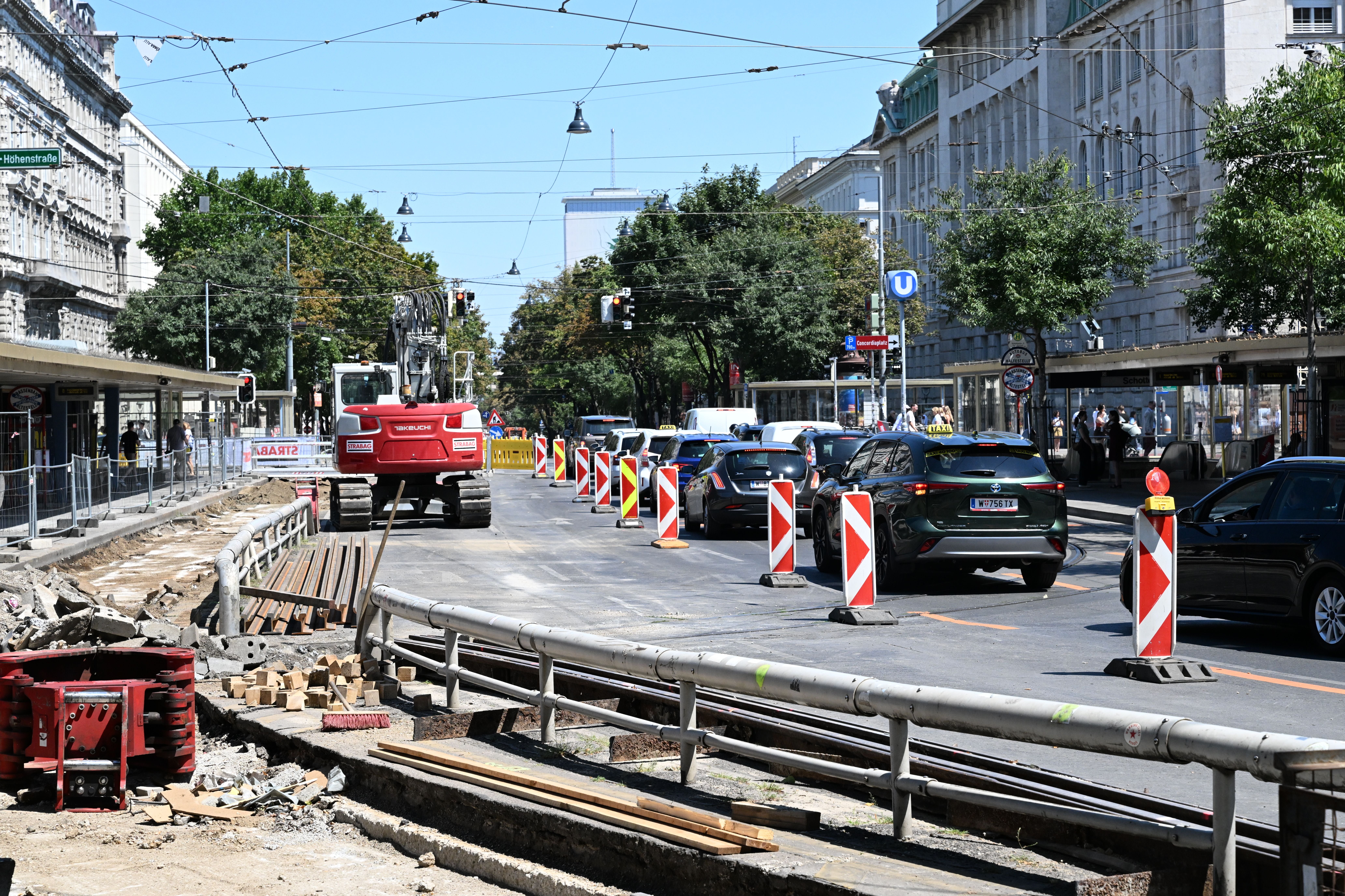 Baustelle mit Bagger und Absperrungen, Autos im Stau, Gebäude und U-Bahn-Zeichen im Hintergrund.