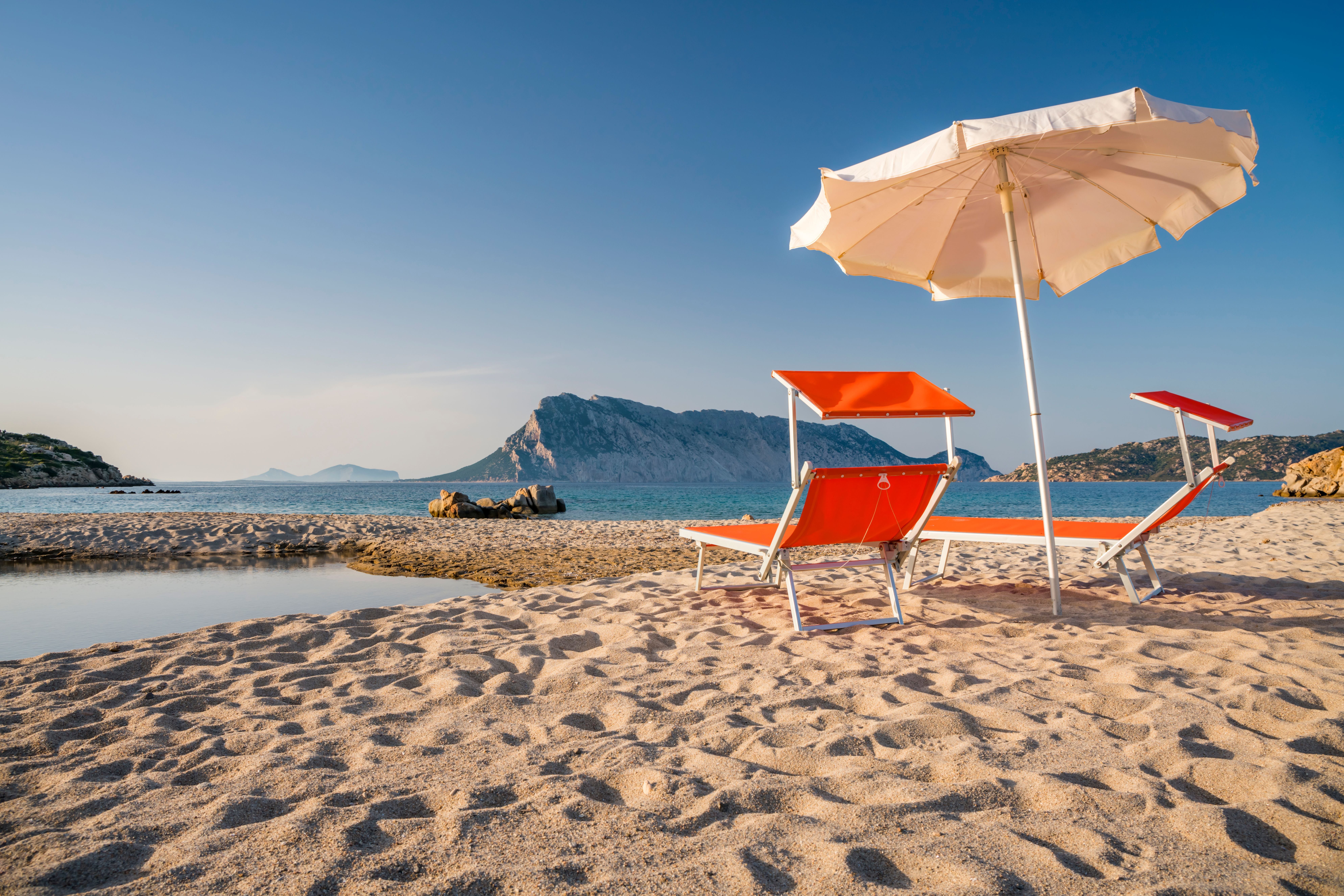 Zwei Sonnenliegen und ein Sonnenschirm am Sandstrand mit Blick auf die Insel Tavolara in Sardinien.