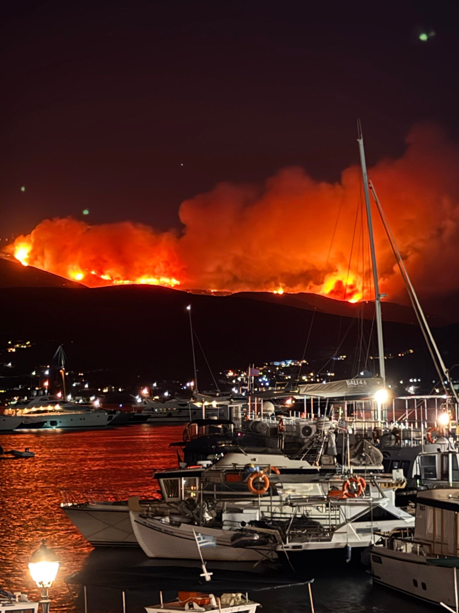 Großes Feuer auf einem Hügel bei Nacht, Boote im Hafen von Paros im Vordergrund.