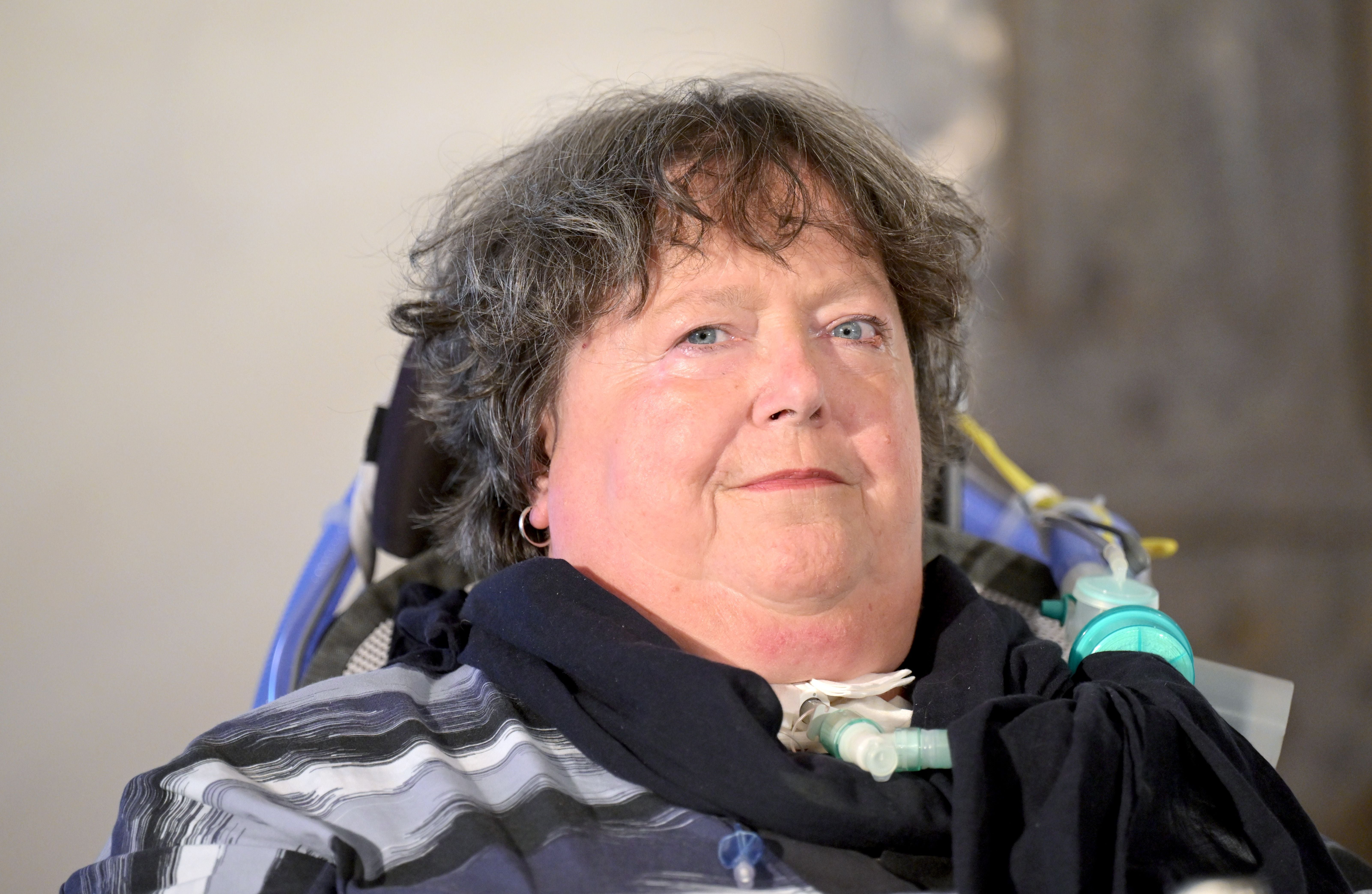 Frau mit Atemhilfe sitzt im Rollstuhl und blickt in die Kamera, unscharfer Hintergrund.