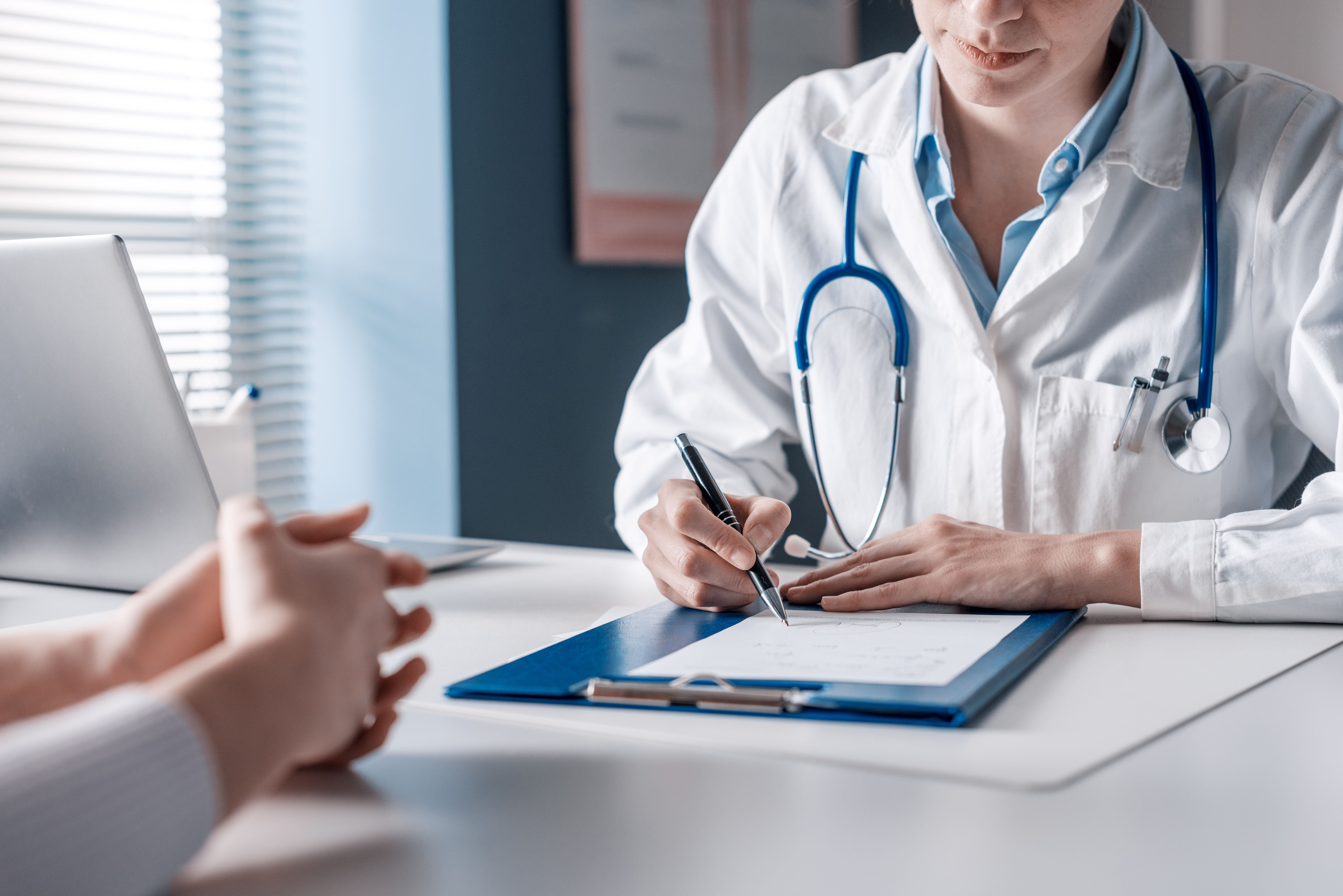 Ärztin schreibt am Schreibtisch ein Rezept, Patient sitzt ihr gegenüber.