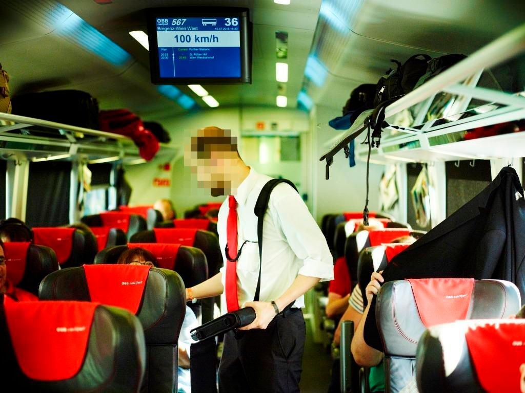 Ein Schaffner kontrolliert Fahrkarten in einem belebten ÖBB-Zugabteil mit roten Sitzbezügen.