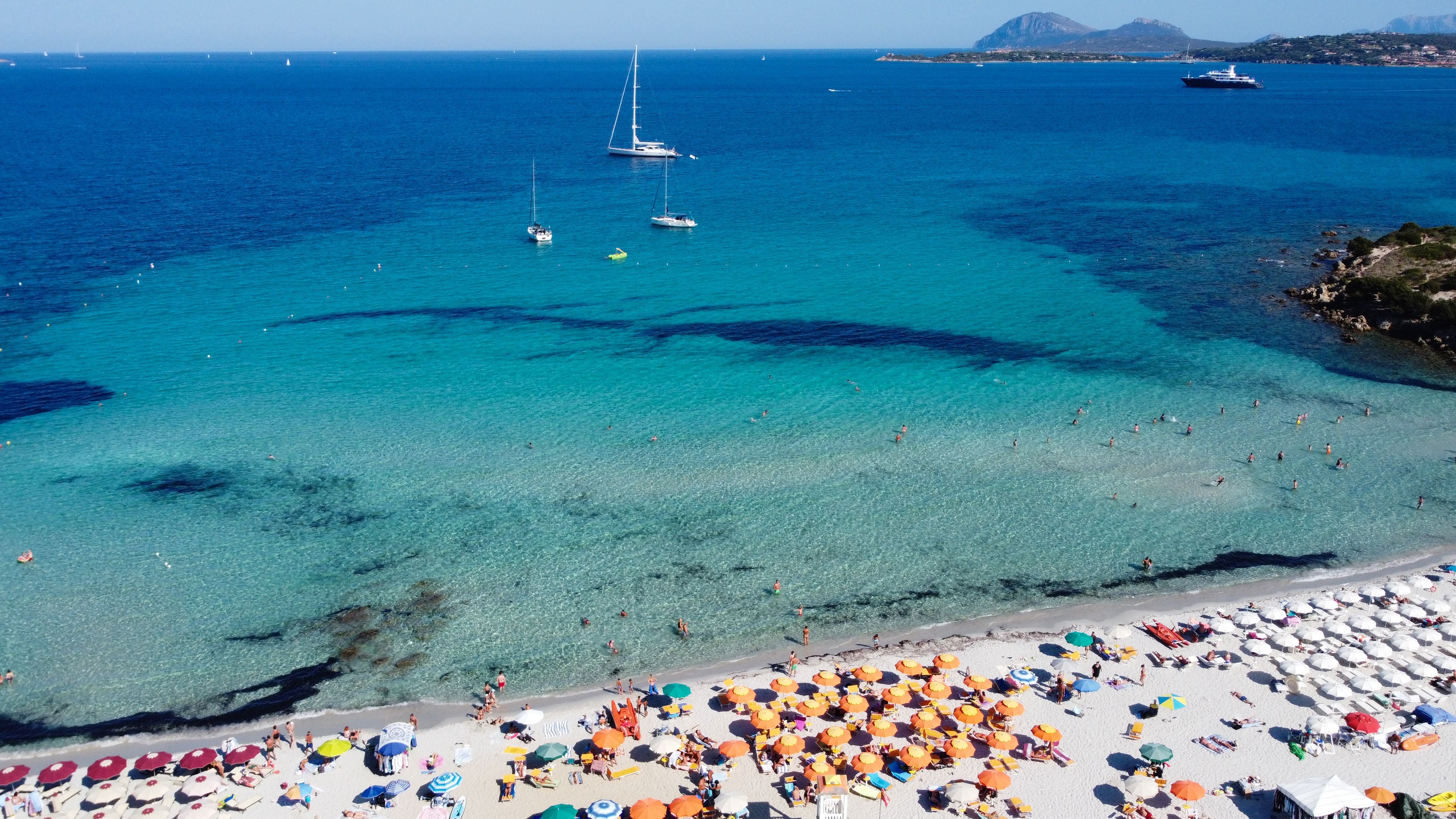 Überfüllter Sandstrand mit bunten Sonnenschirmen, türkisfarbenem Meer und Booten im Hintergrund.