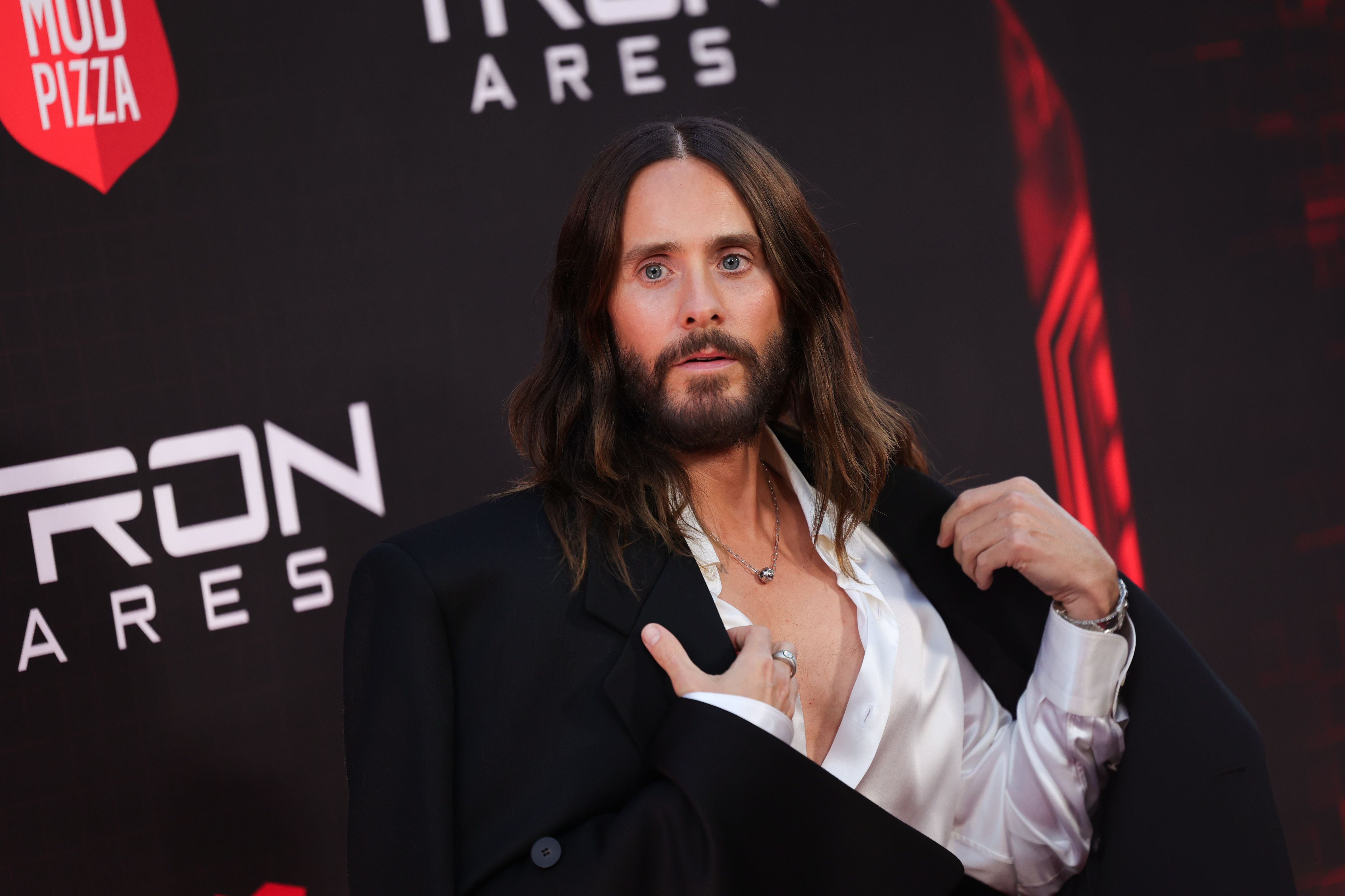 Eine Person mit langen Haaren und Bart posiert im Anzug vor einem "Tron: Ares"-Premieren-Hintergrund.