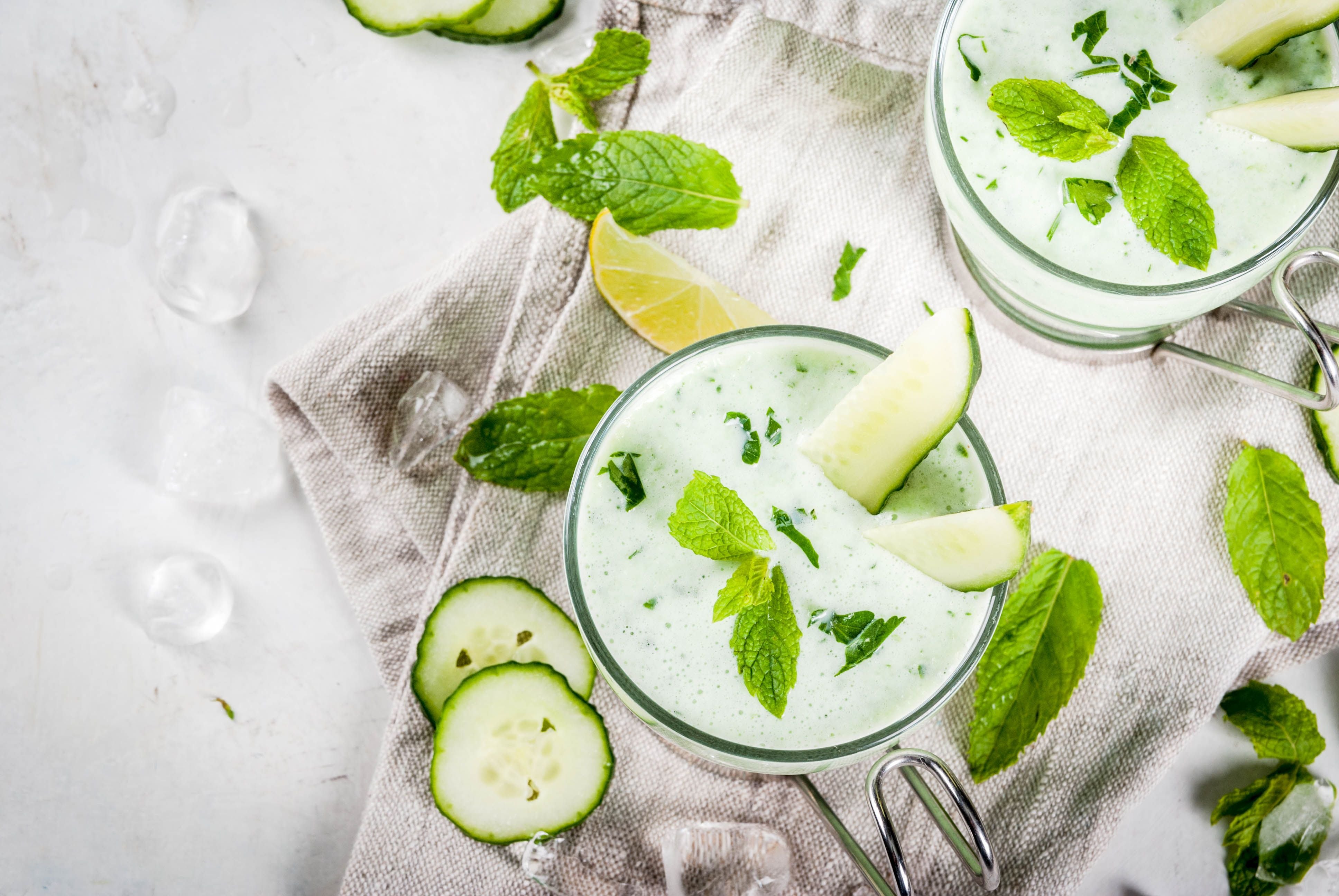 Zwei Gläser kalte Gurken-Avocado-Suppe mit Minze, Gurkenscheiben und Limetten auf weißem Untergrund.