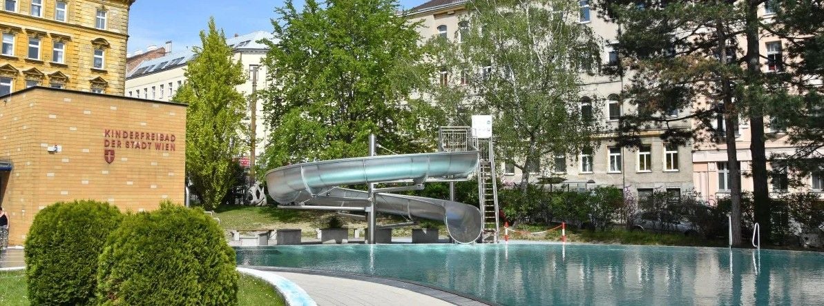Kinderfreibad mit Rutsche und Becken, umgeben von Bäumen und Wohnhäusern in Wien.