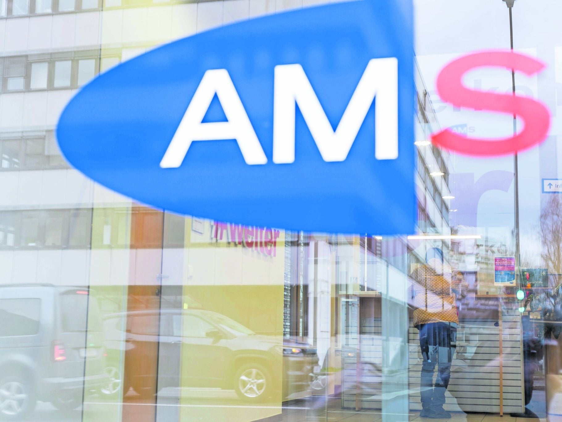 AMS-Logo an einer Glasscheibe mit Spiegelung einer Straße und einer Person im Hintergrund.