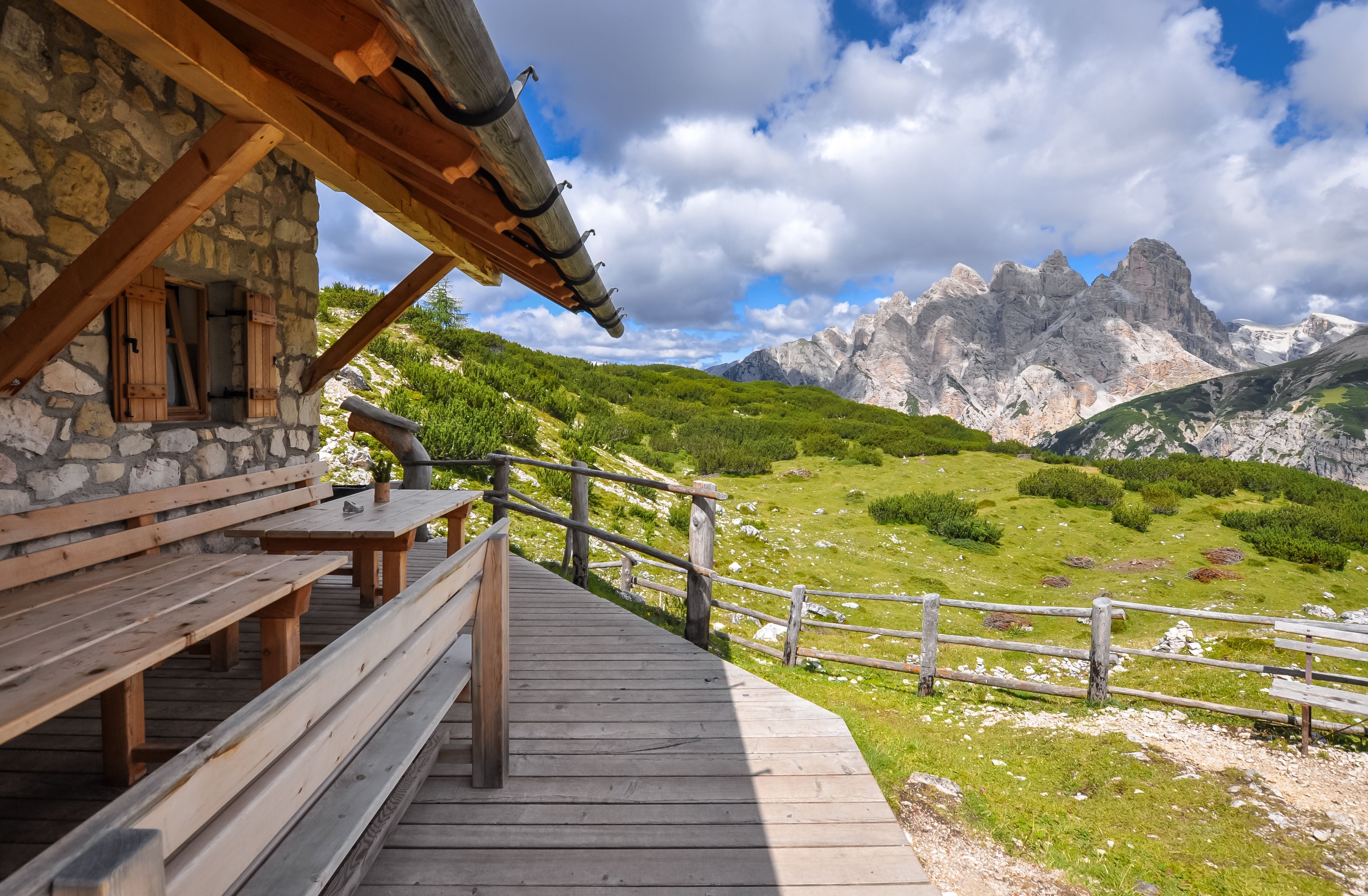 Holzhütte auf einer Blumenwiese vor steilen Bergen, eine Kuh steht im Hintergrund.