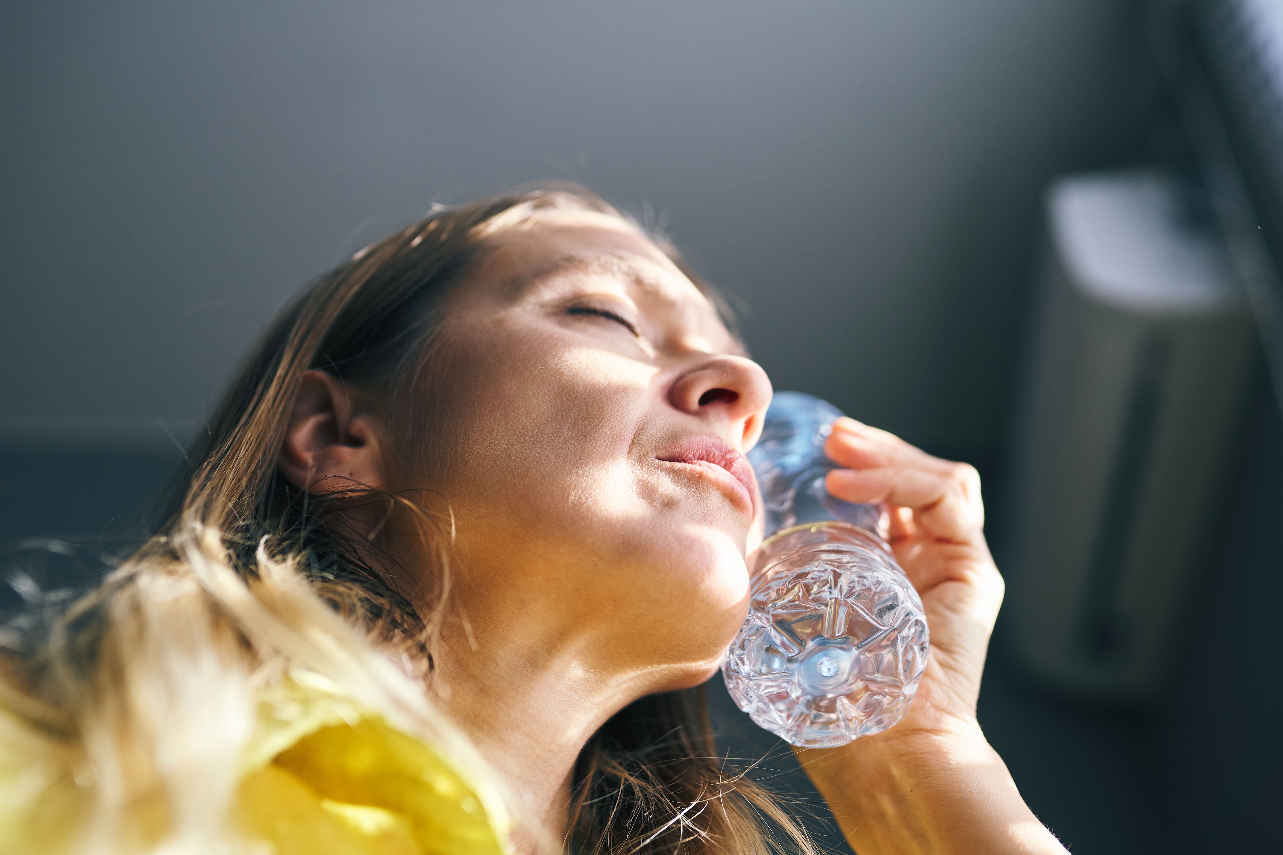 Frau kühlt sich mit einer Wasserflasche am Hals ab, Sonnenlicht betont Überhitzung und Unwohlsein.
