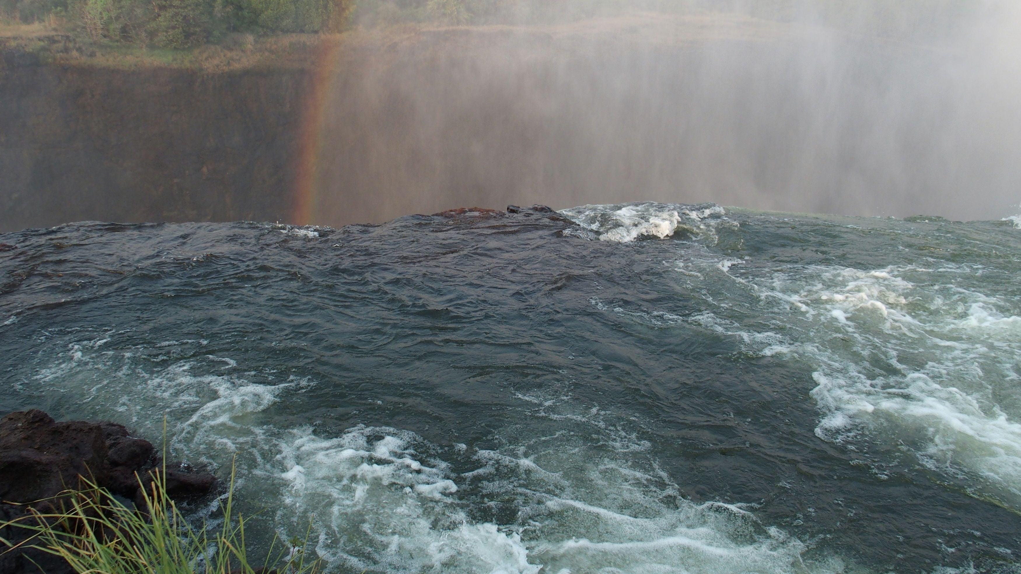 Blick auf Devil’s Pool am Rand der Victoriafälle mit Wasserströmung und Regenbogen im Nebel.