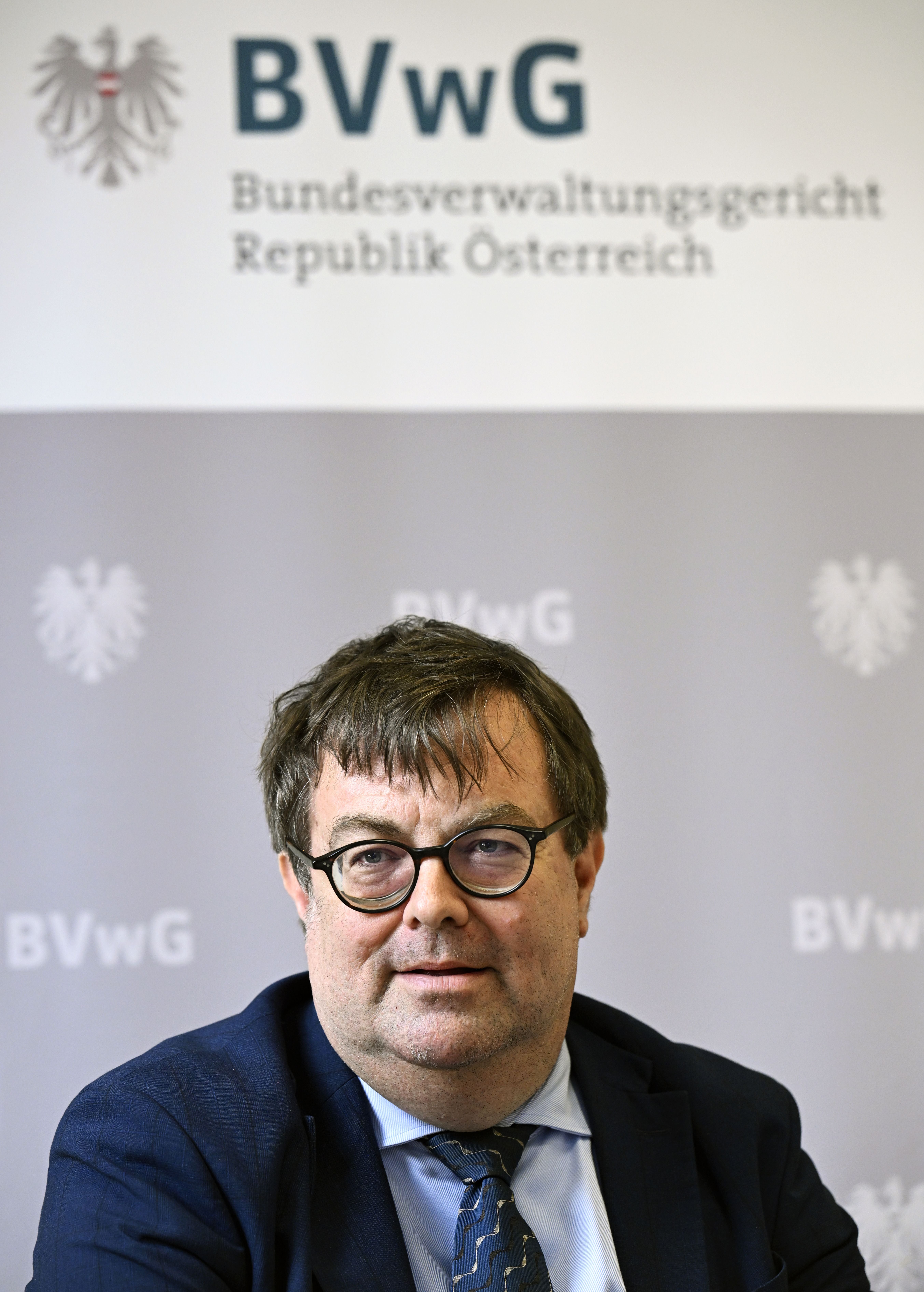 Ein Mann mit Brille und Anzug sitzt vor einem Hintergrund des Bundesverwaltungsgerichts Österreich.