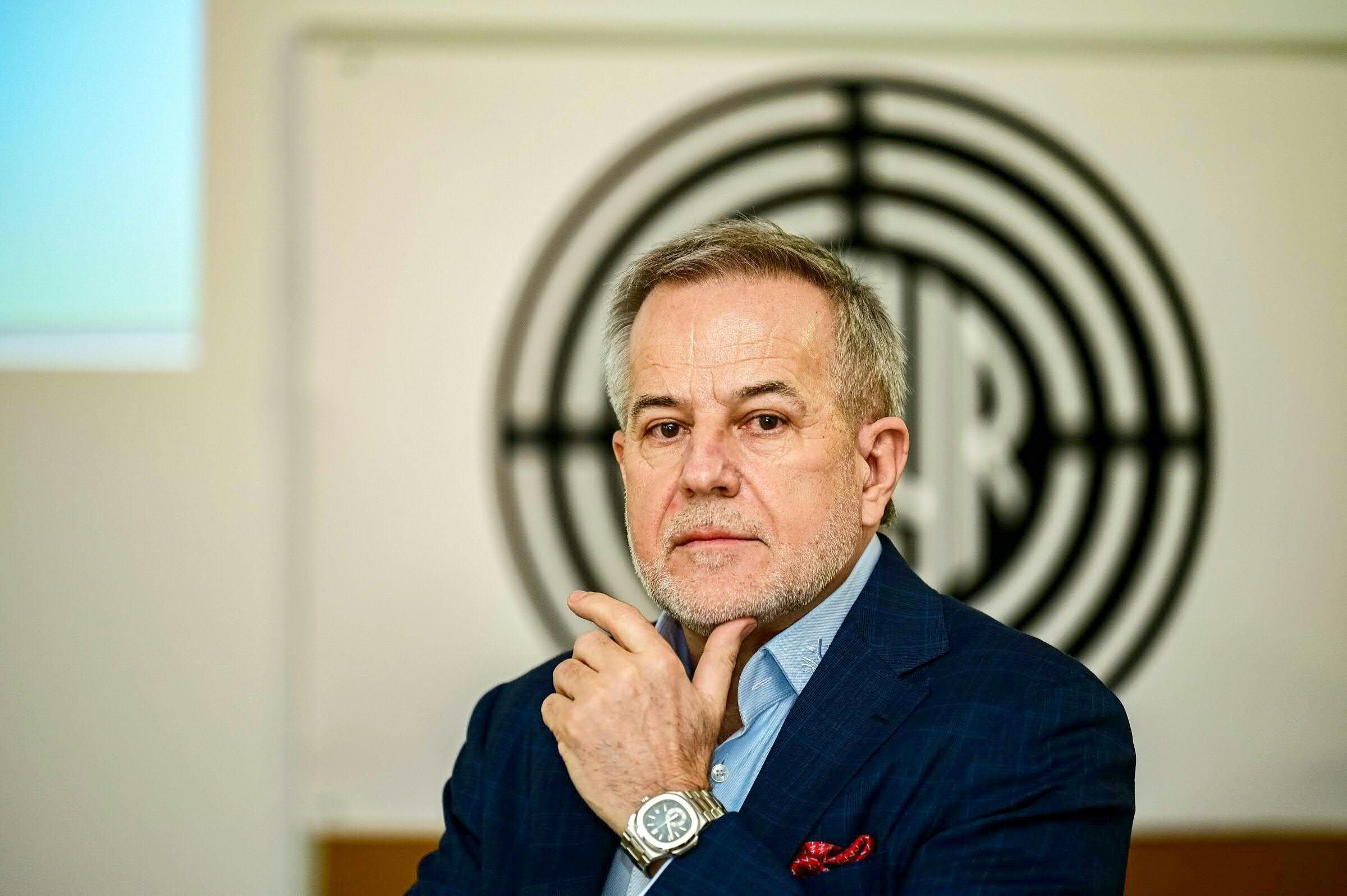 Ein Mann im Anzug mit Uhr posiert nachdenklich vor einem schwarz-weißen Logo im Hintergrund.