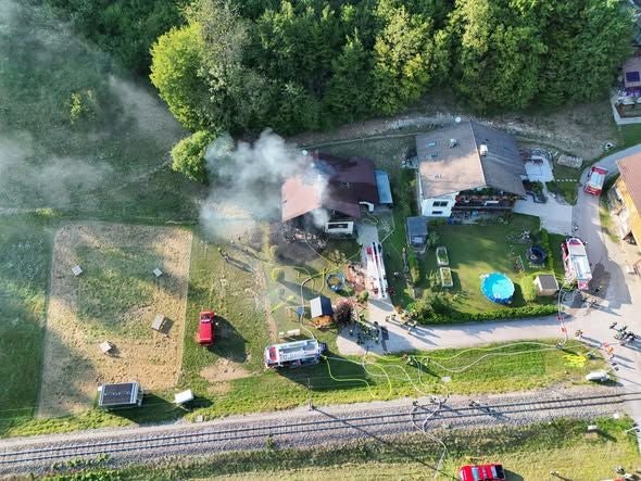 Rauch steigt aus einem brennenden Haus; Feuerwehrfahrzeuge und Einsatzkräfte sind vor Ort.
