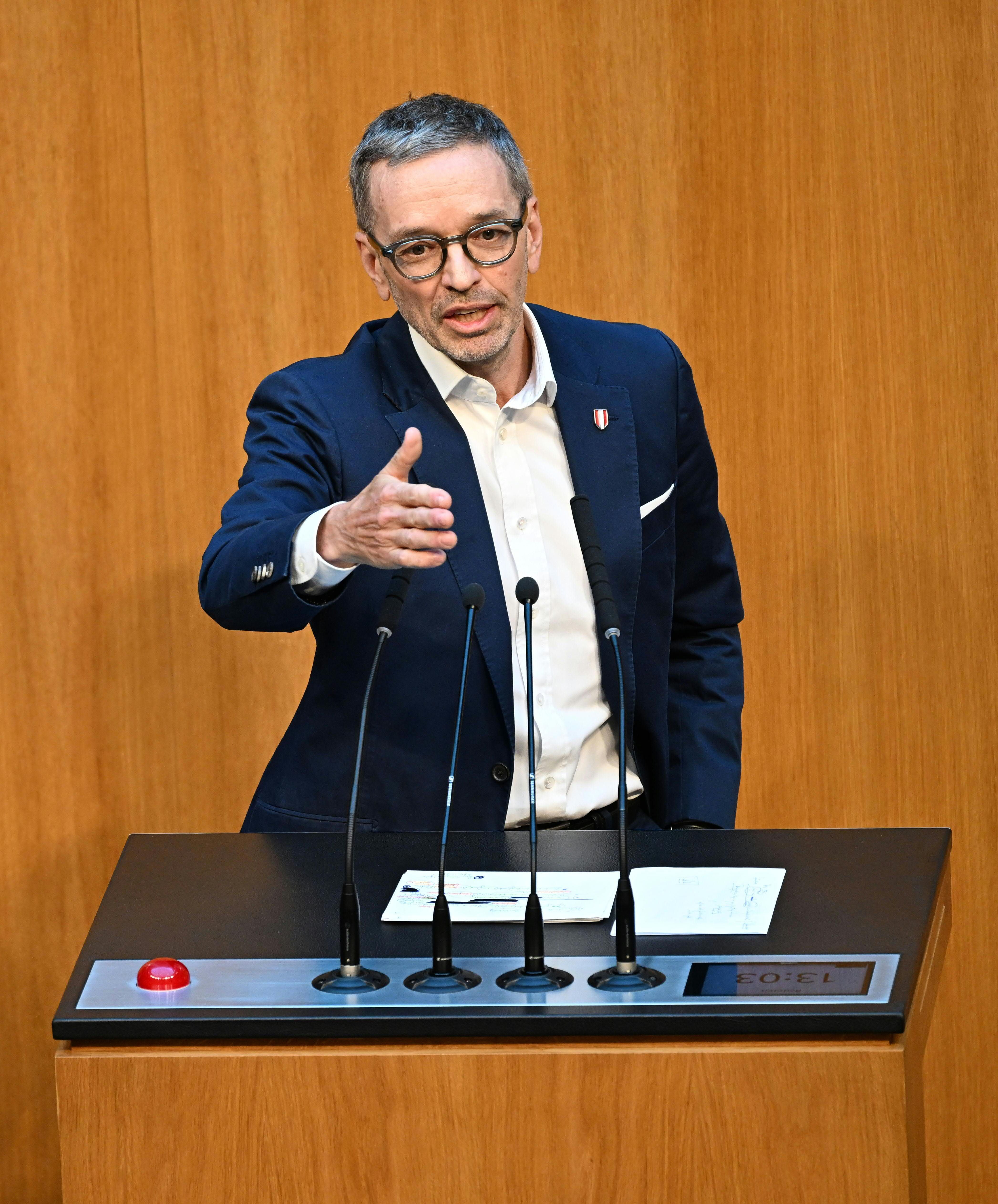 Ein Mann im Anzug spricht an einem Rednerpult mit vier Mikrofonen im Parlament.