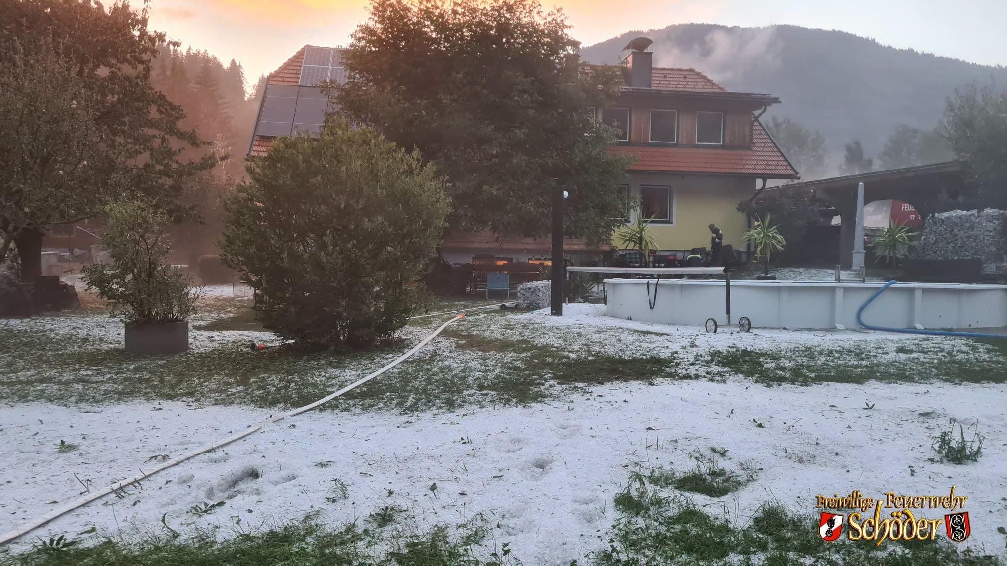 Ein Garten und Haus mit Hagel bedeckt, Bäume, Pool und Feuerwehr im Hintergrund, Steiermark.