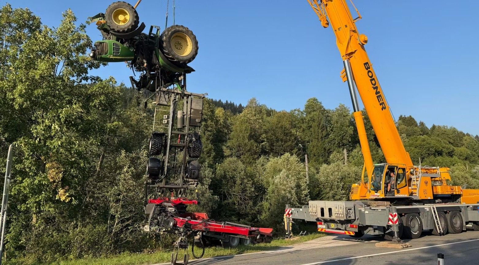 Ein gelber Kran hebt einen umgestürzten Traktor samt Anhänger an einer Landstraße.