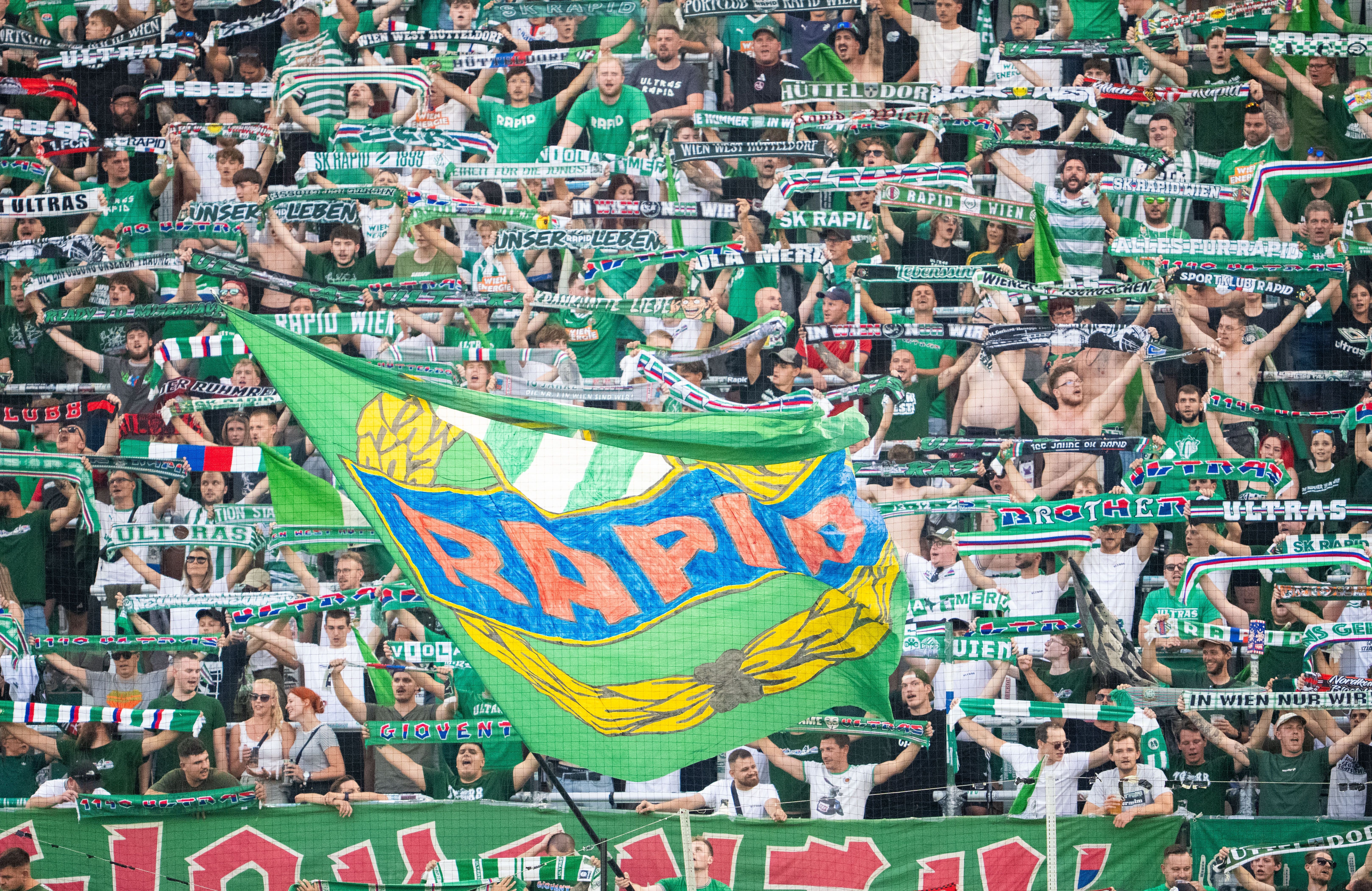 SK Rapid Fans jubeln mit grünen Schals und großer Rapid-Flagge im Fußballstadion.