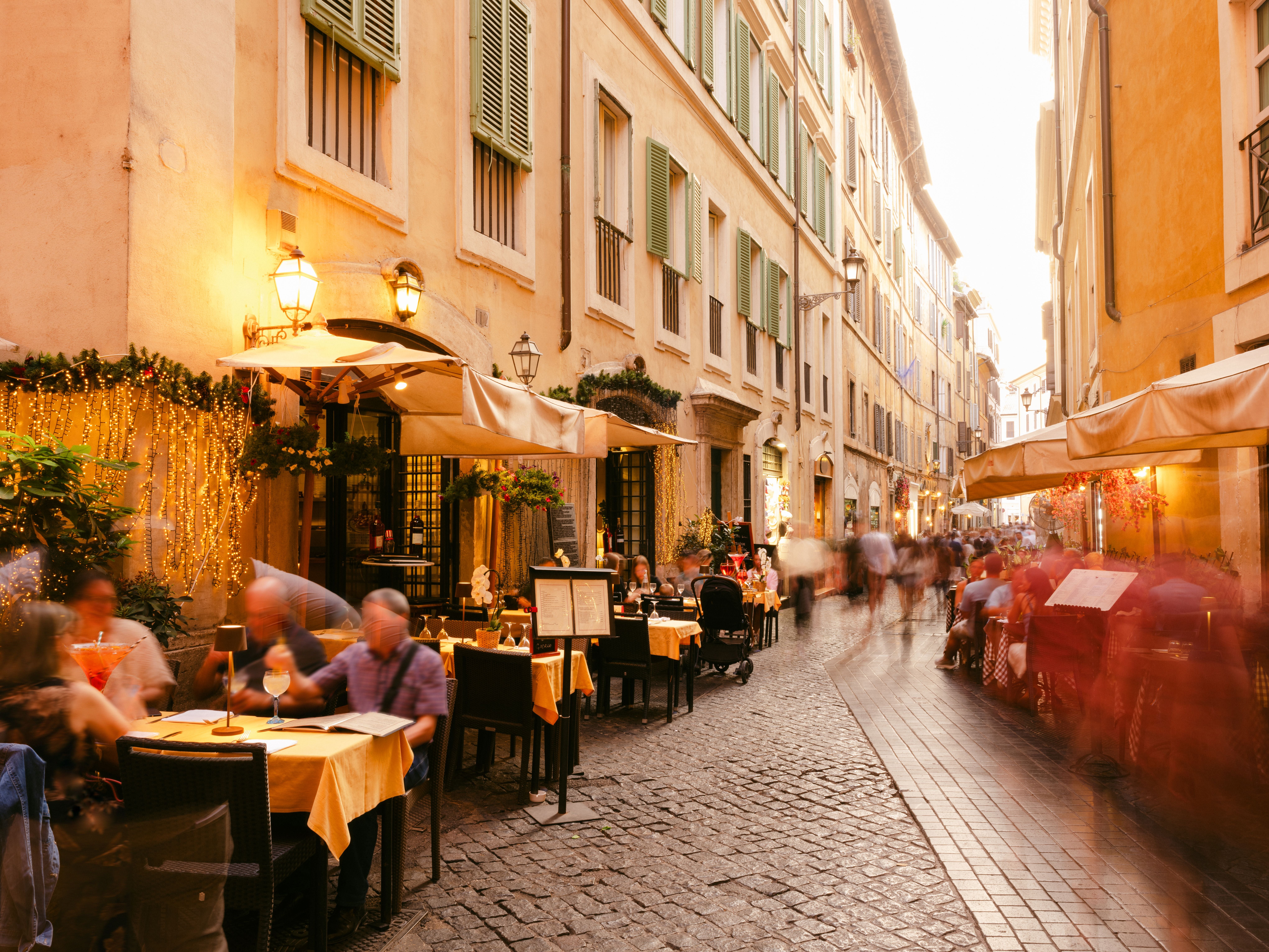 Menschen essen abends auf einer kopfsteingepflasterten Straße in Rom im Freien vor Restaurants.