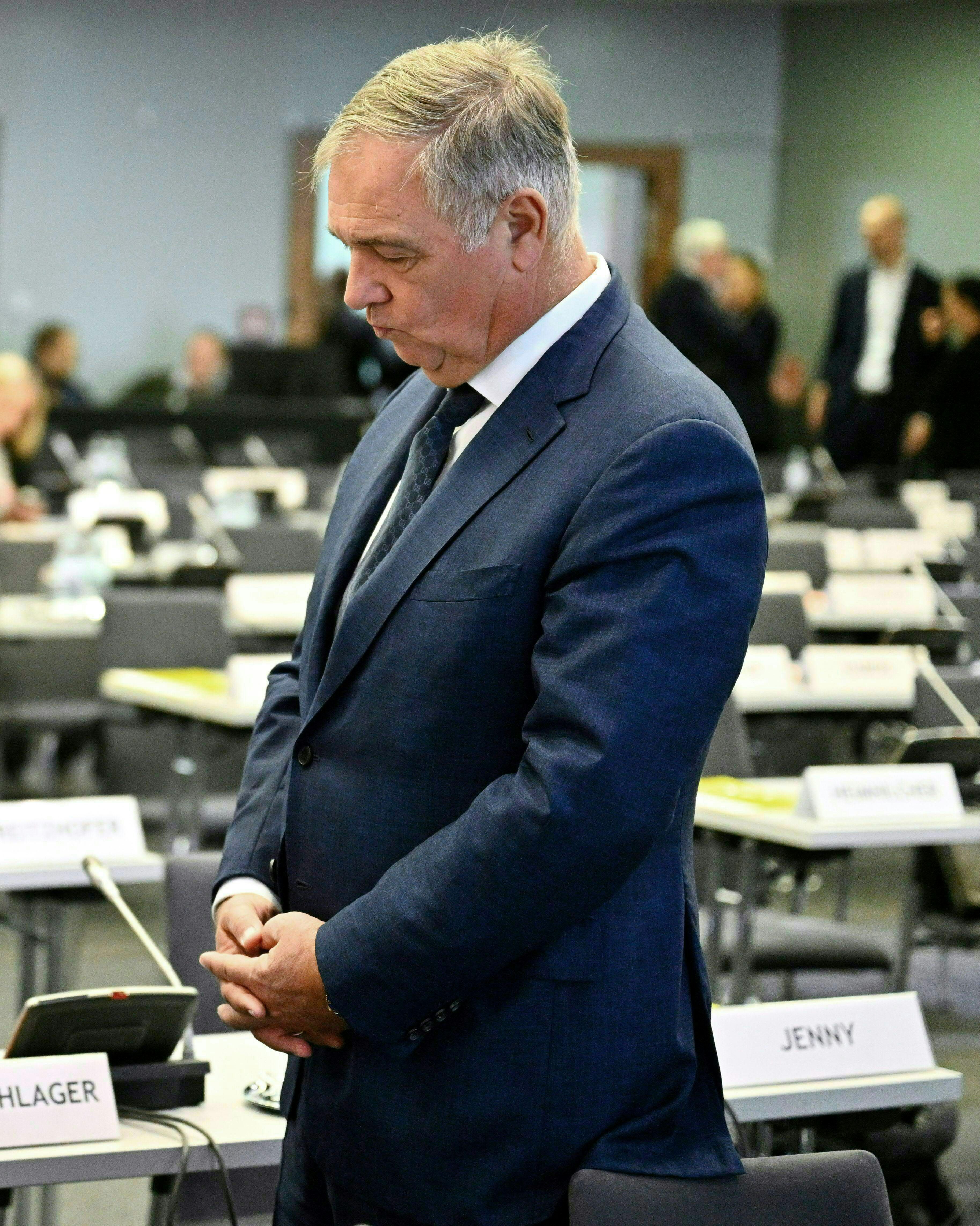 Ein Mann im Anzug steht nachdenklich in einem Konferenzraum mit Namensschildern auf den Tischen.