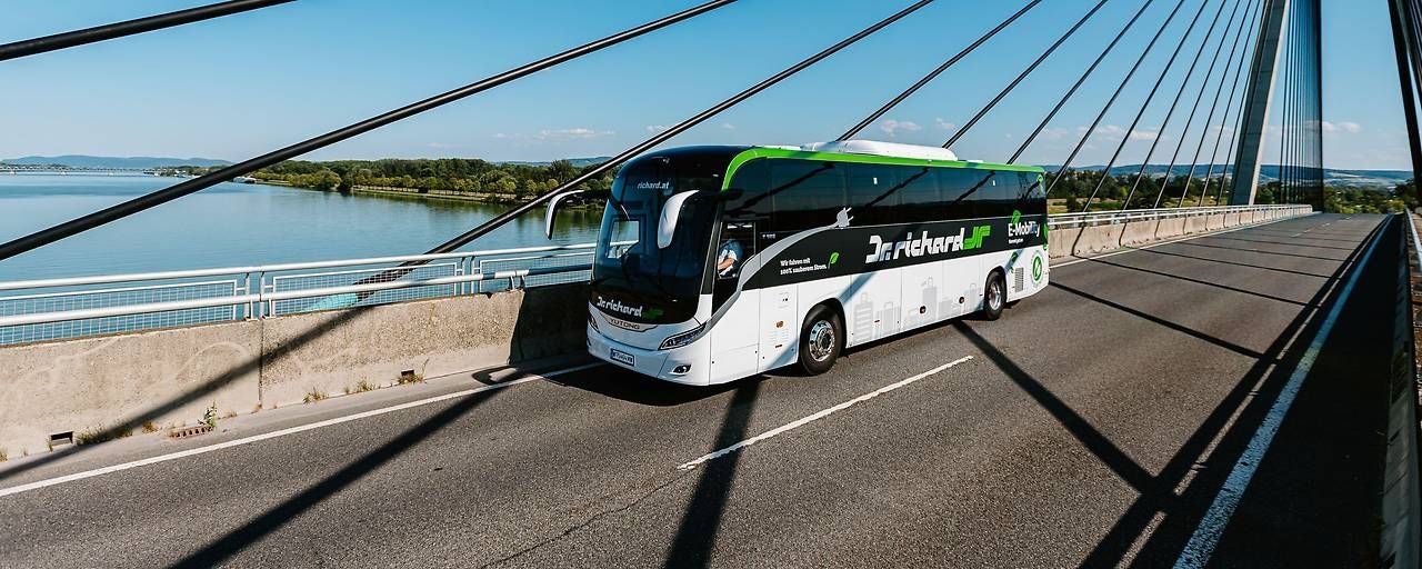 Ein moderner E-Reisebus fährt über eine Brücke mit Fluss im Hintergrund bei sonnigem Wetter.
