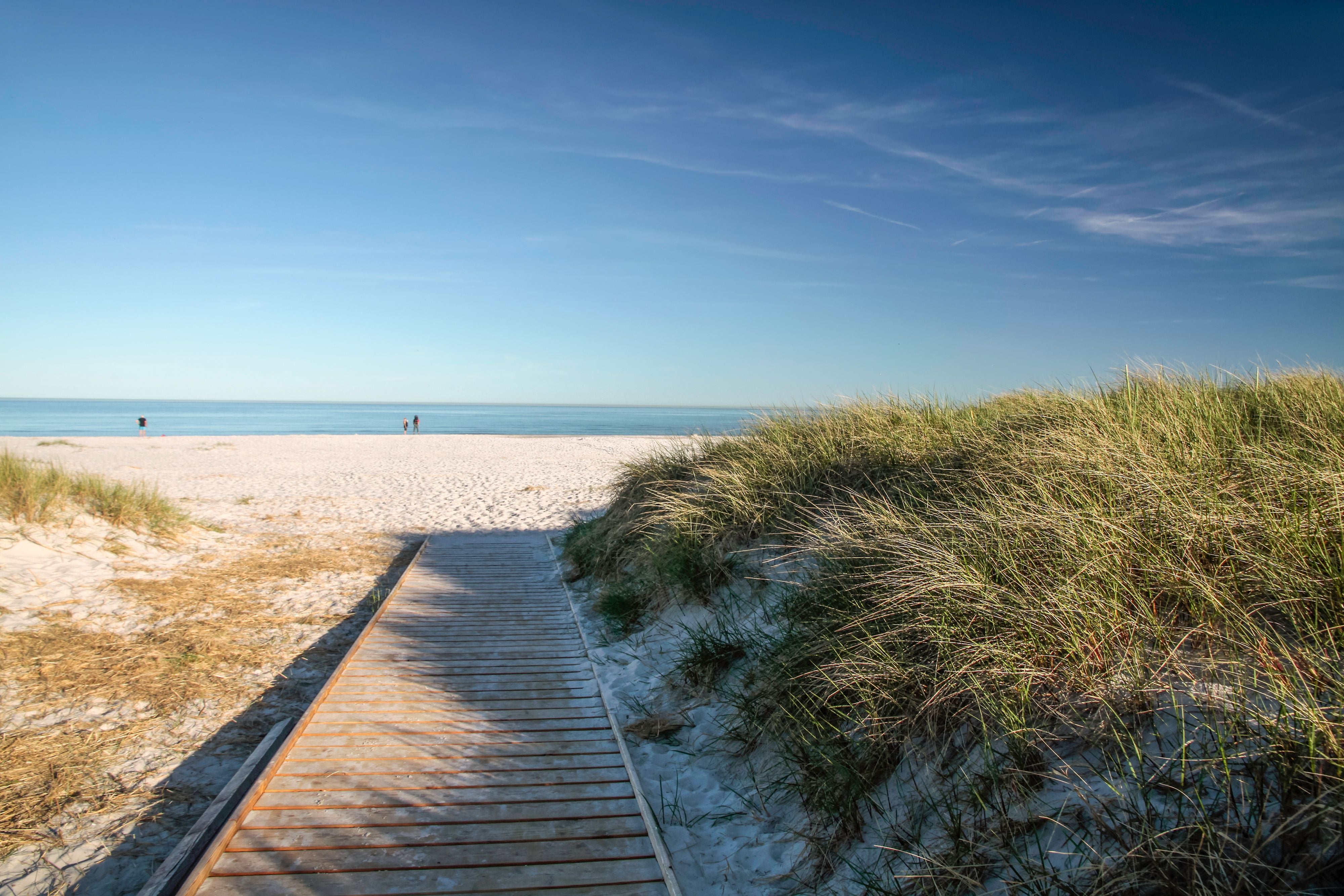 Holzsteg führt durch Dünen zu einem weißen Sandstrand mit Blick auf das ruhige Meer und blauen Himmel.