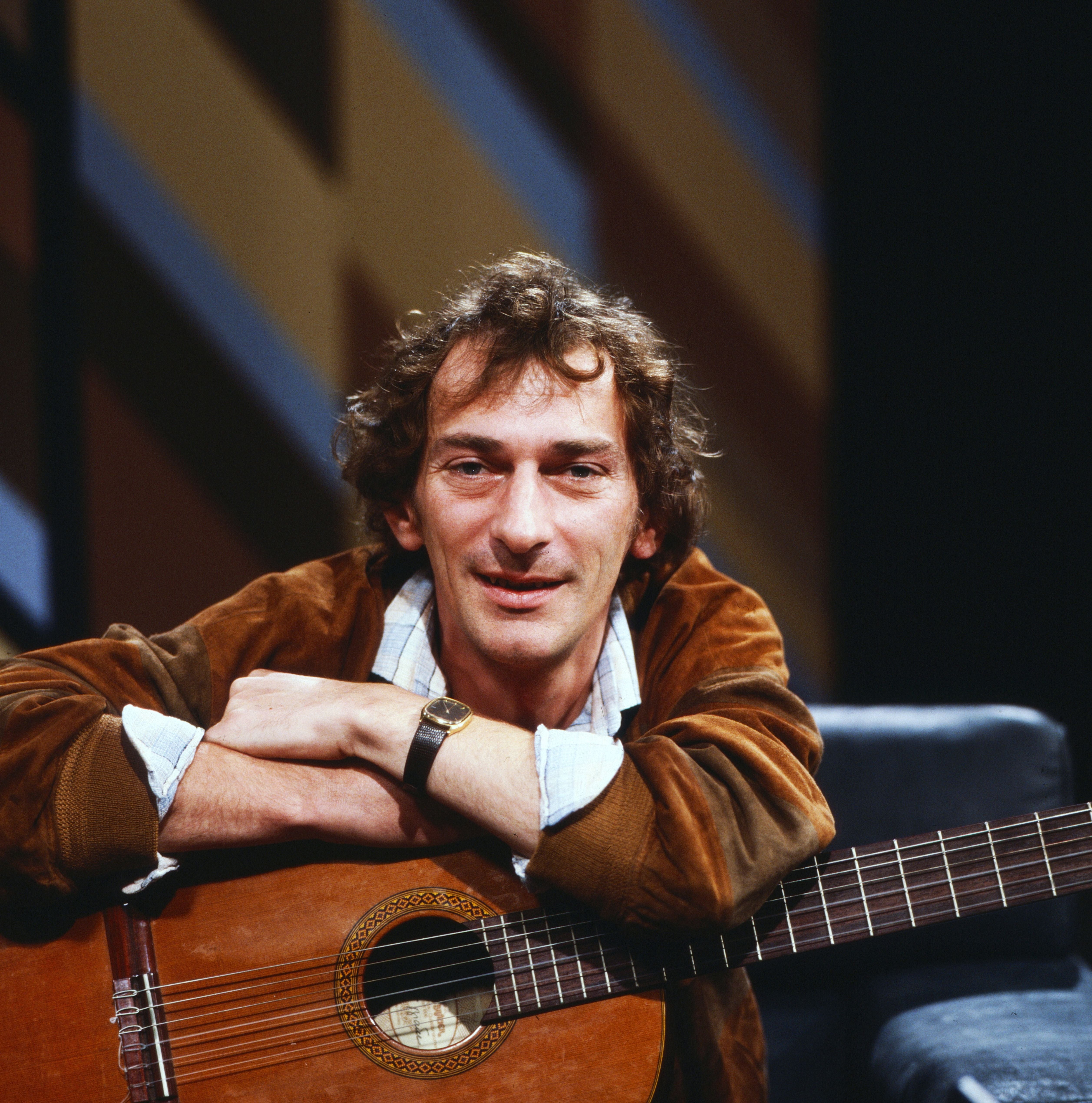 Mann mit Gitarre sitzt entspannt, stützt die Arme auf dem Instrument und lächelt in die Kamera.