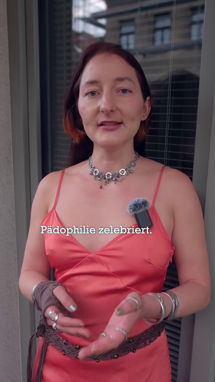 Frau mit roten Haaren und orangem Kleid spricht, Text: „Pädophilie zelebriert.“