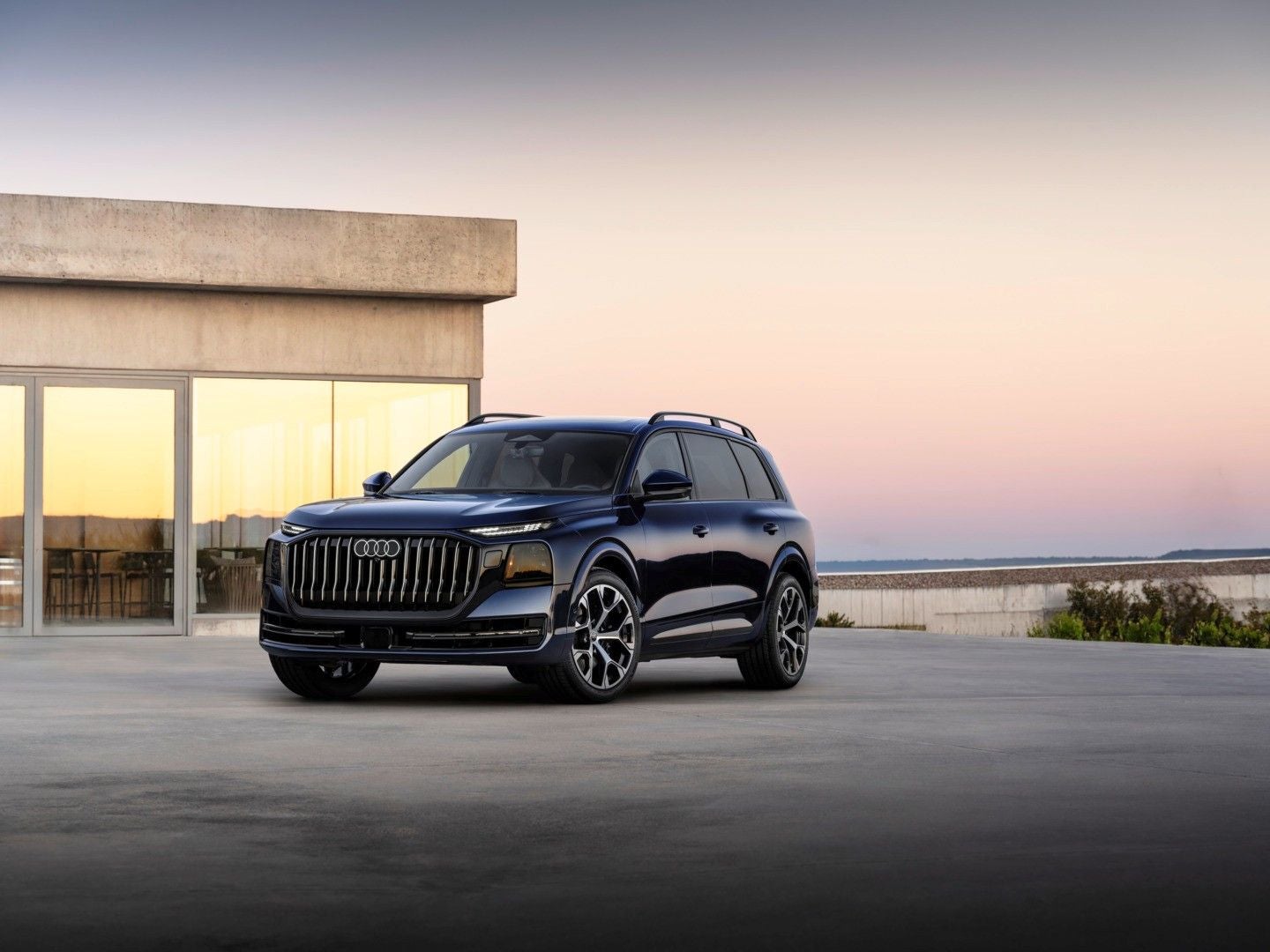 Ein dunkelblauer Audi Q9 SUV steht vor einem modernen Gebäude bei Sonnenuntergang.