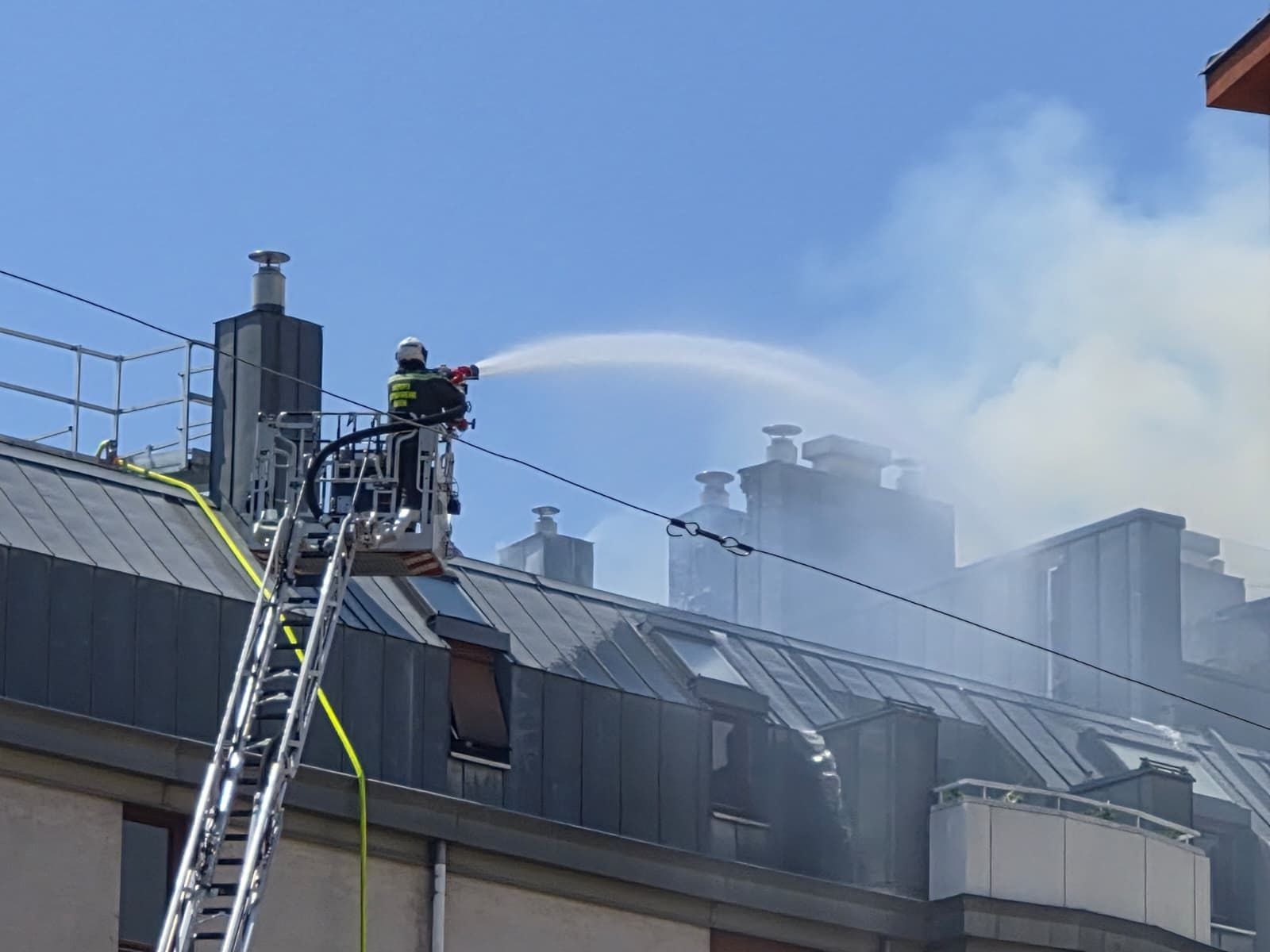 Feuerwehr löscht mit Wasserstrahl einen Dachstuhlbrand in Meidling, Rauch steigt auf.