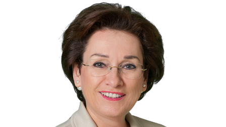 Porträt einer lächelnden Frau mit Brille und kurzen braunen Haaren vor neutralem Hintergrund.