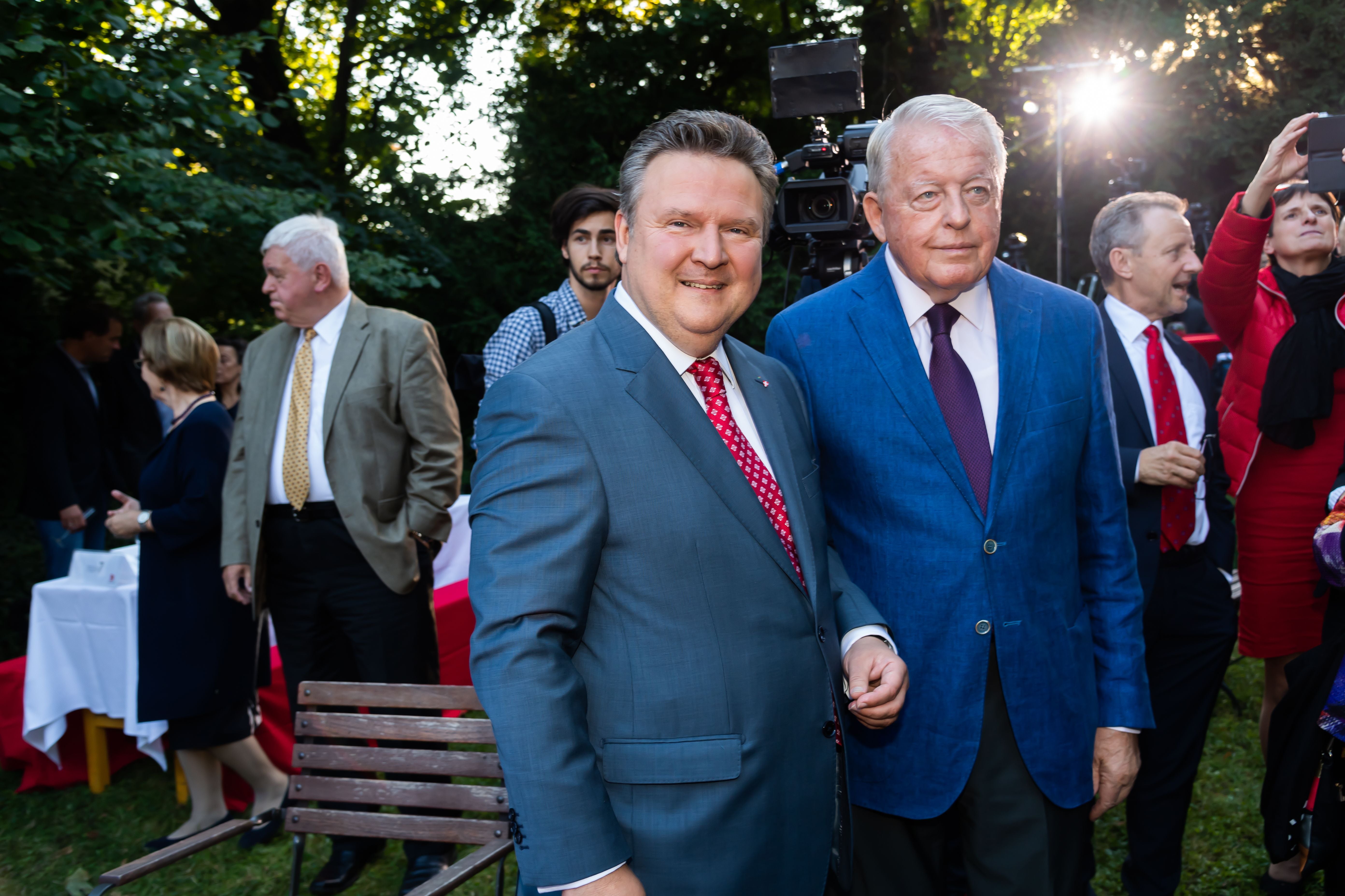 Zwei Männer im Anzug posieren bei einer Feier im Freien, umgeben von weiteren Gästen.