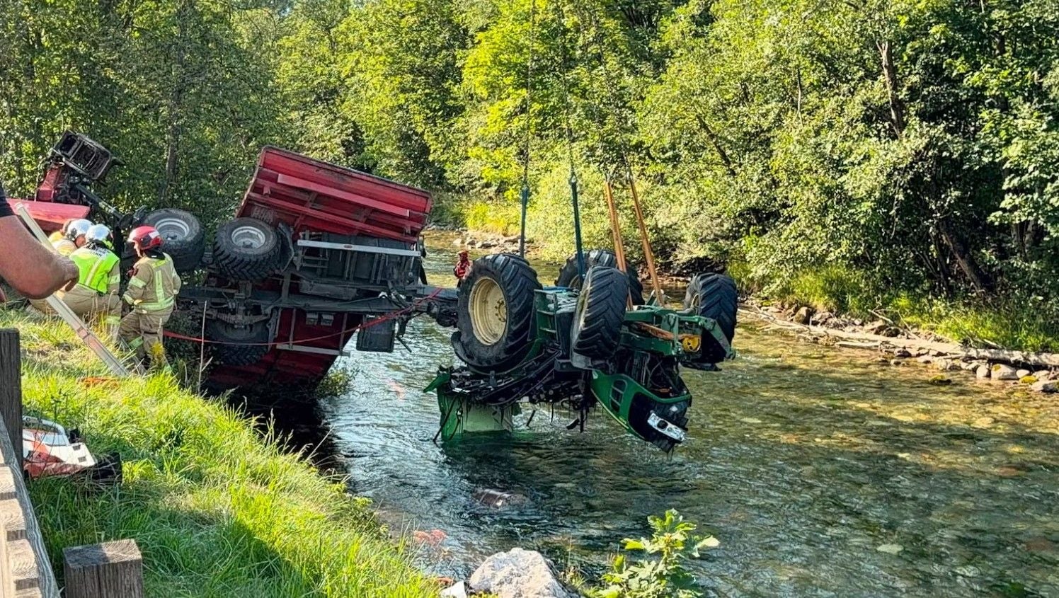 Feuerwehr zieht umgestürzten Traktor und Anhänger aus einem Bach in bewaldeter Umgebung.