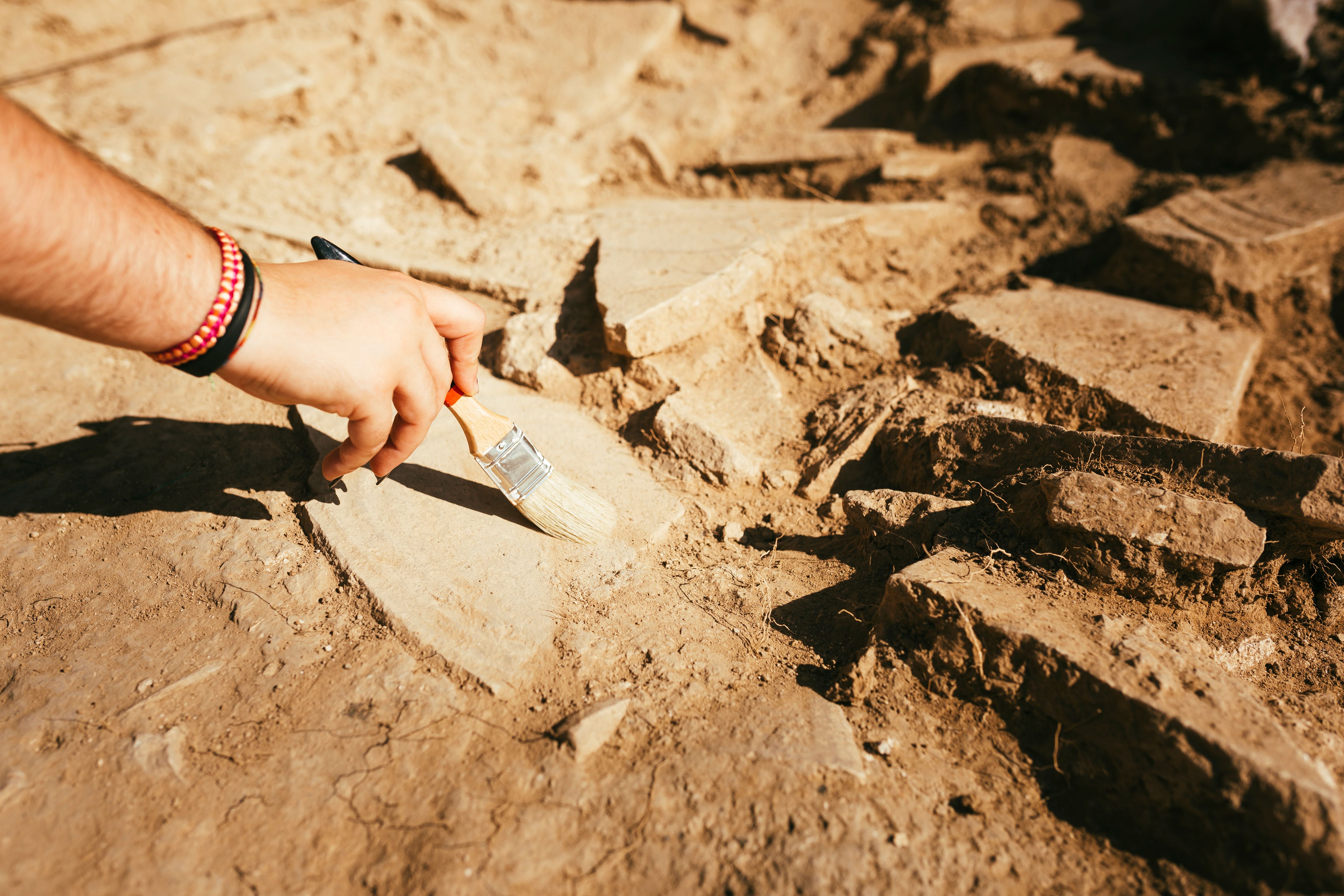 Hand mit Pinsel entfernt vorsichtig Staub von Keramikstücken an einer archäologischen Ausgrabungsstätte.