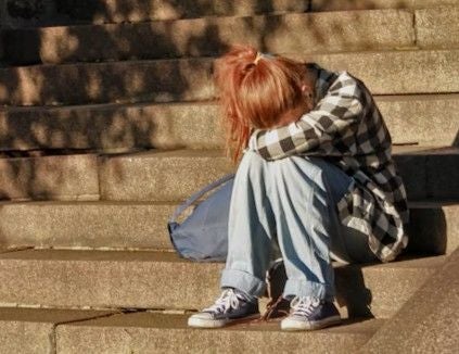 Ein Mädchen sitzt traurig auf Treppen, lehnt den Kopf auf die Arme und hat eine Tasche daneben.