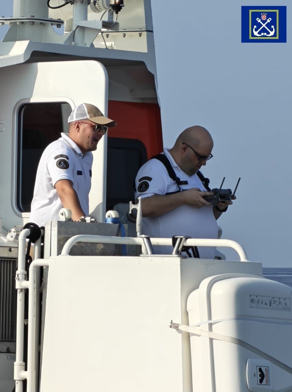Zwei Männer in Uniform bedienen ein Gerät auf dem Deck eines Bootes mit Marineemblem.