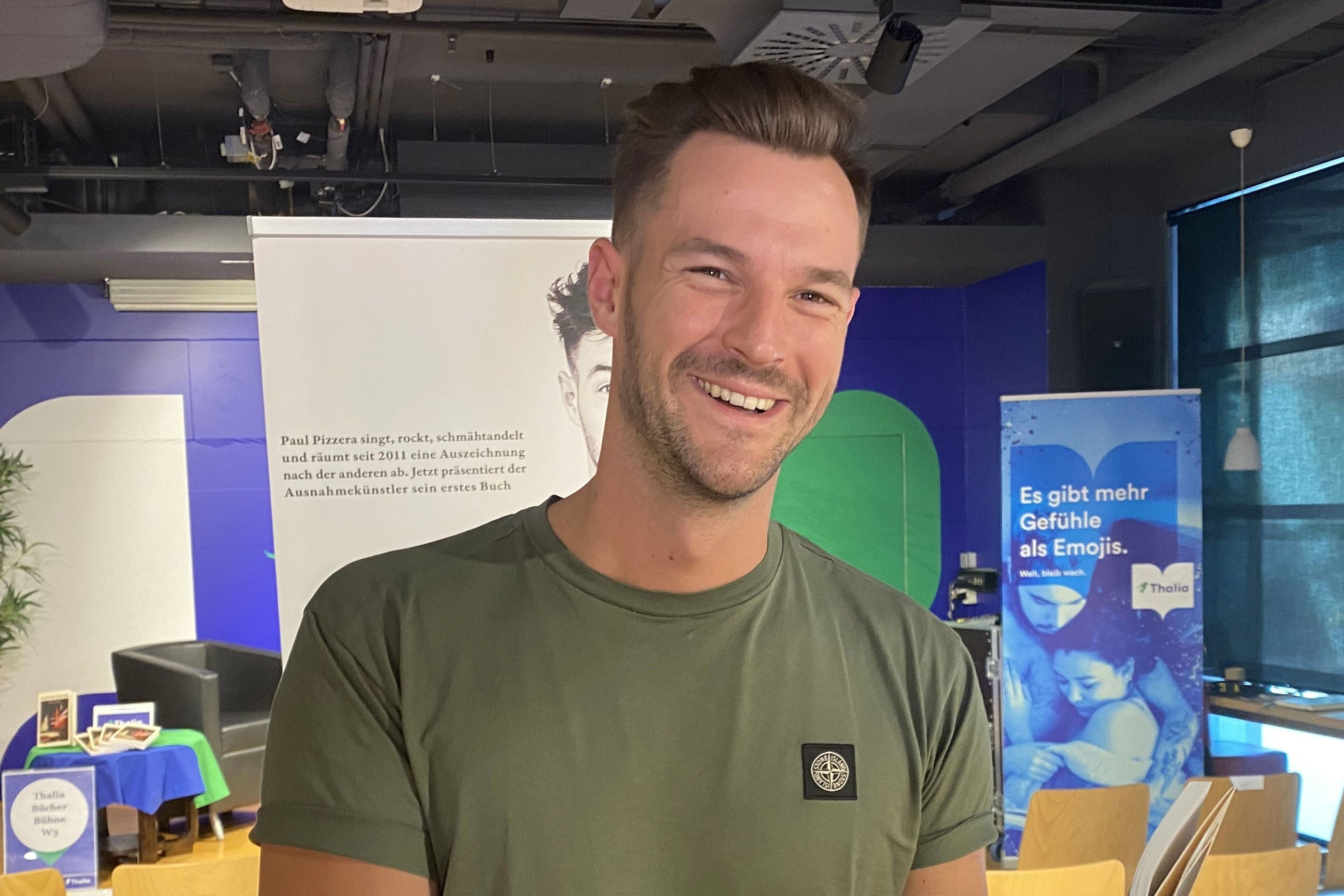 Junger Mann mit grünem T-Shirt lächelt in einem modernen Veranstaltungsraum in die Kamera.