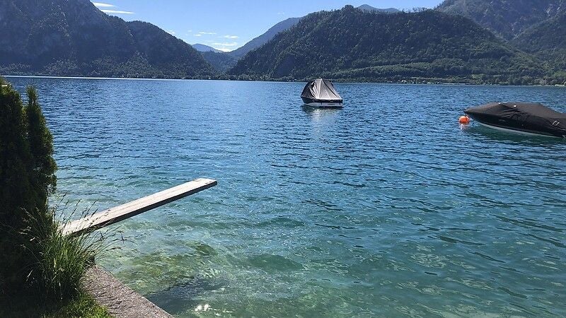 Blick auf einen klaren See mit Sprungbrett, zwei abgedeckten Booten und Bergen im Hintergrund.
